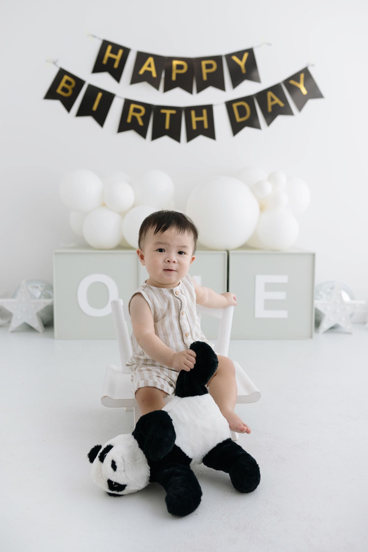 Child sitting on panda plush toy with birthday decorations including banner, balloons and blocks in background.