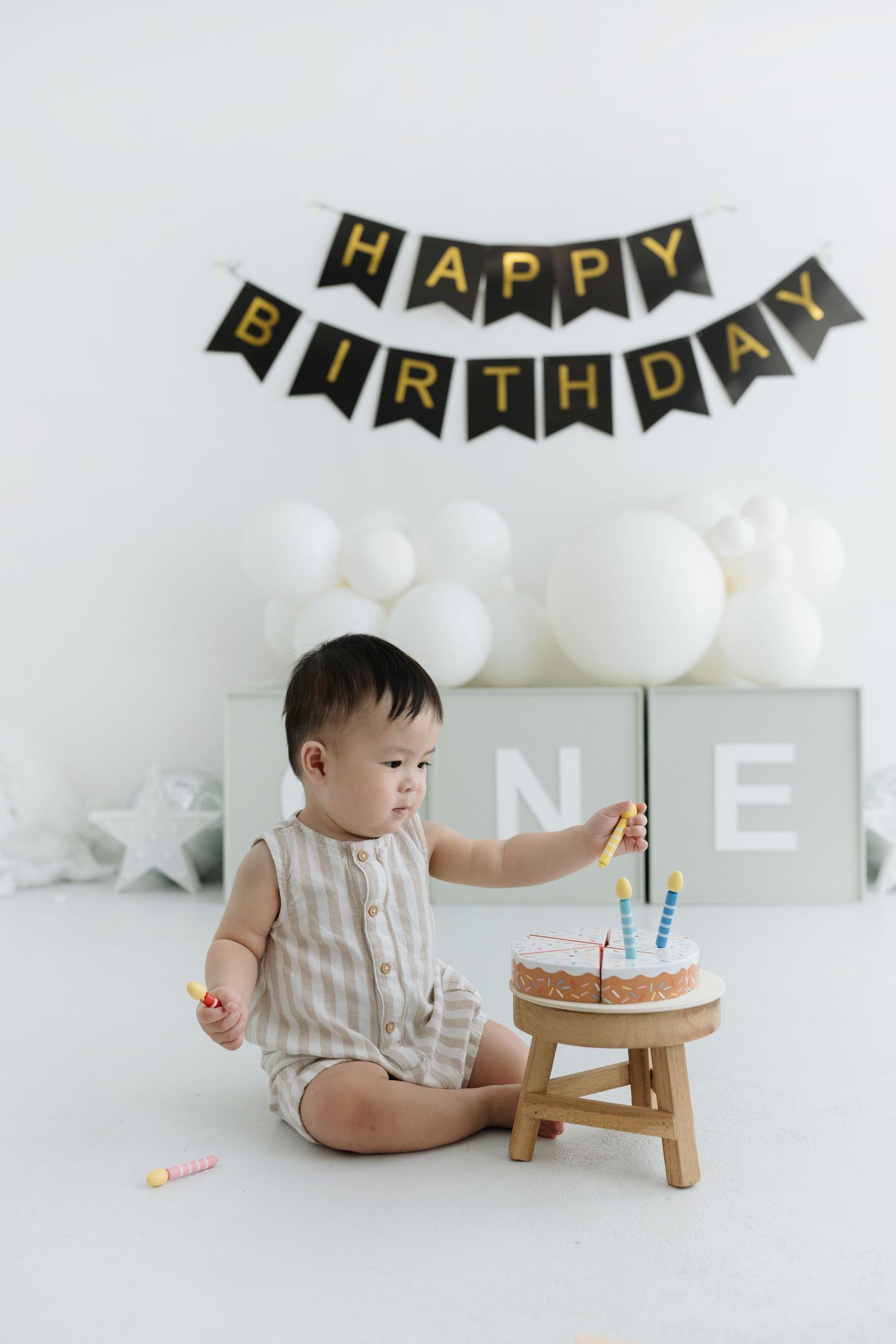 A first birthday celebration setup with black and gold banner, white balloons, and small wooden cake stand against white backdrop.