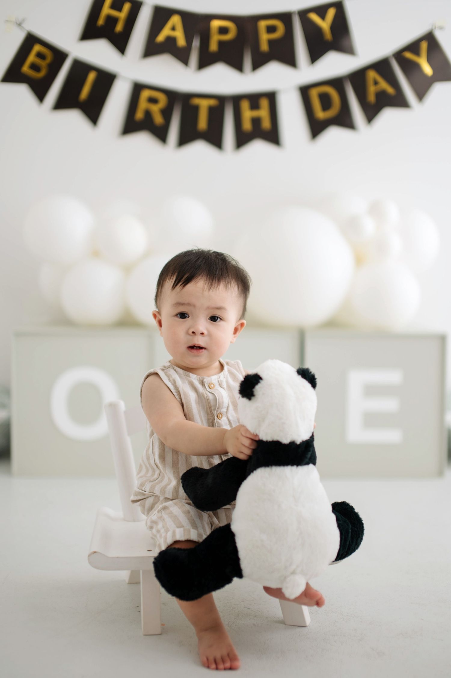Baby in white outfit plays with black and white panda plush toy under happy birthday banner and white balloons.