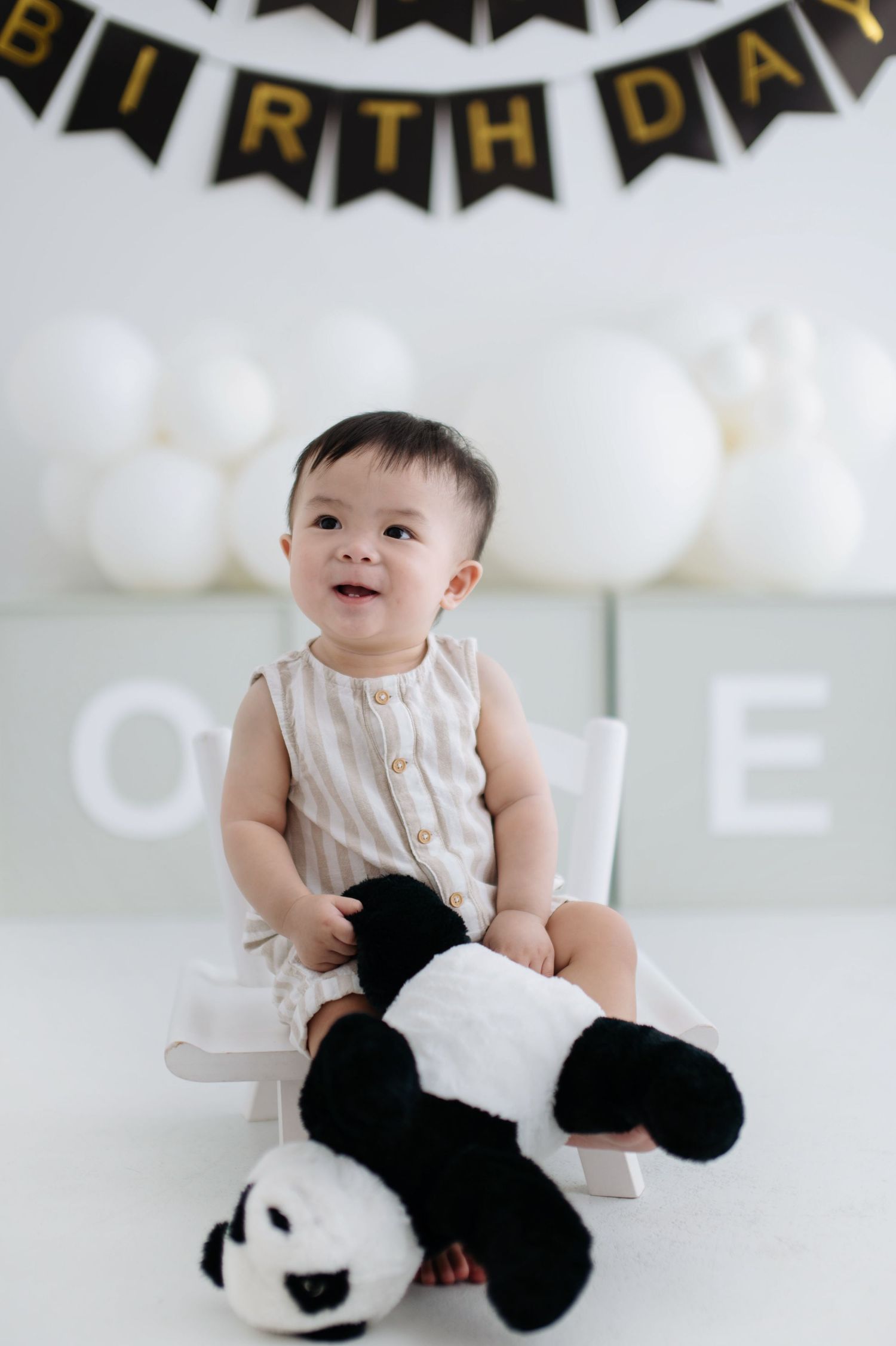 Smiling baby sits in white chair with panda stuffed animal under birthday banner and balloon decorations.