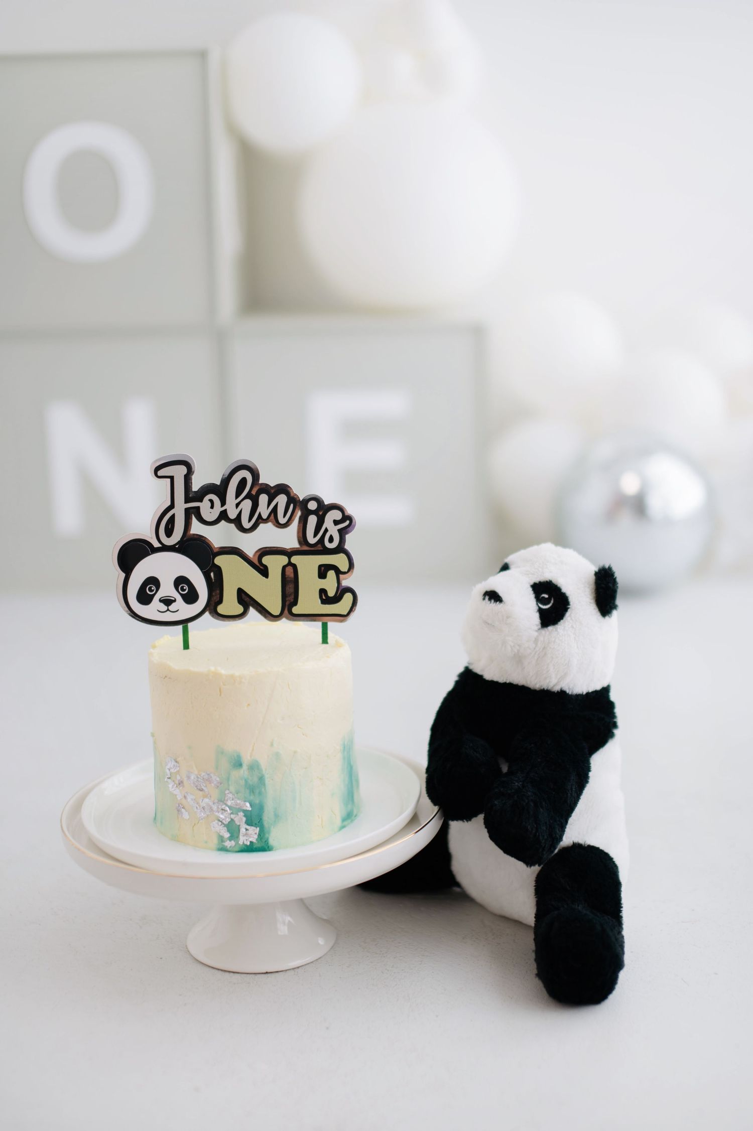 A white birthday cake with black panda decorations and 'ONE' cake topper sits on a cake stand against a light background.