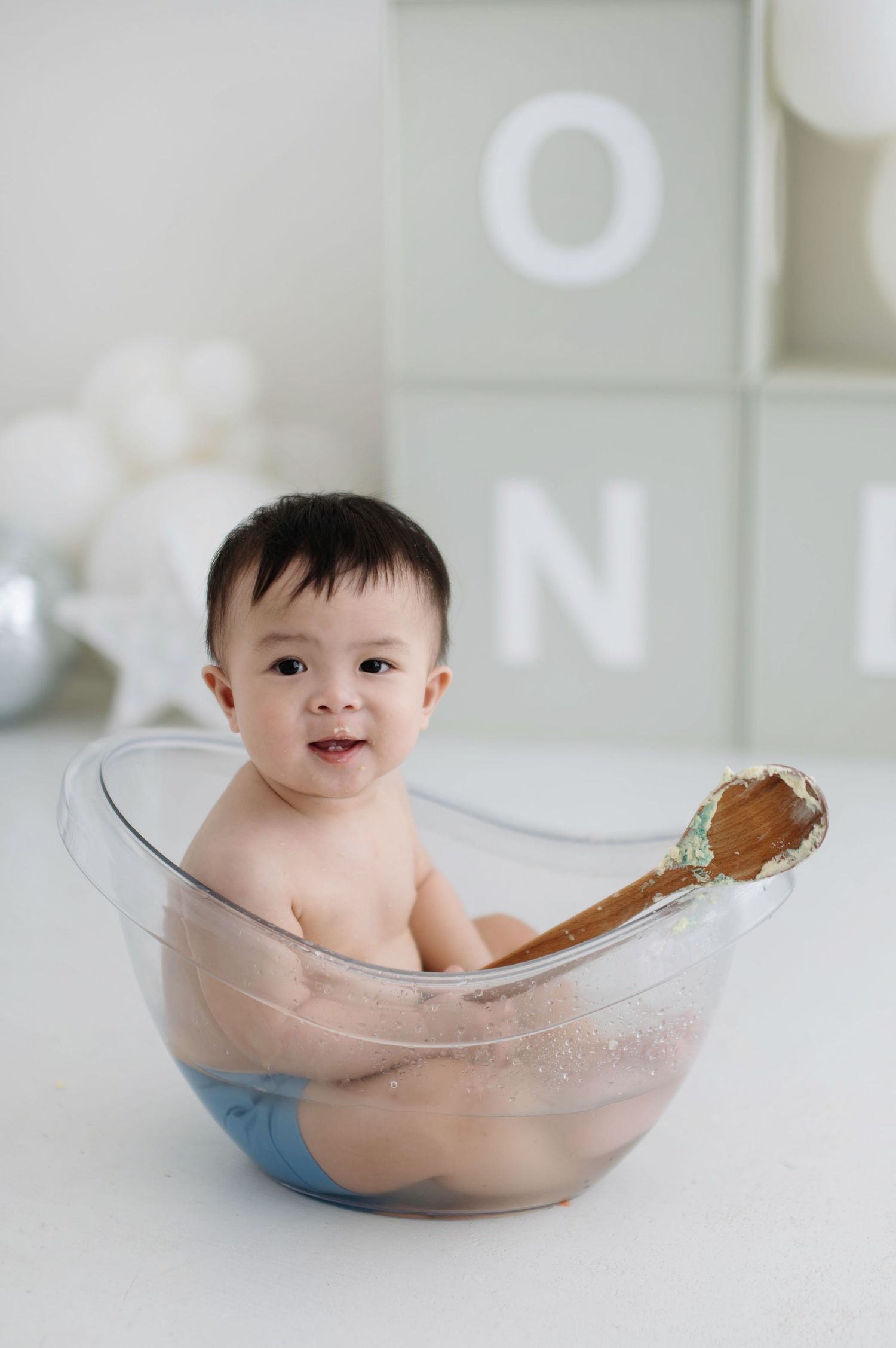 Baby sitting in clear glass mixing bowl during bath time with white wall decor in background.