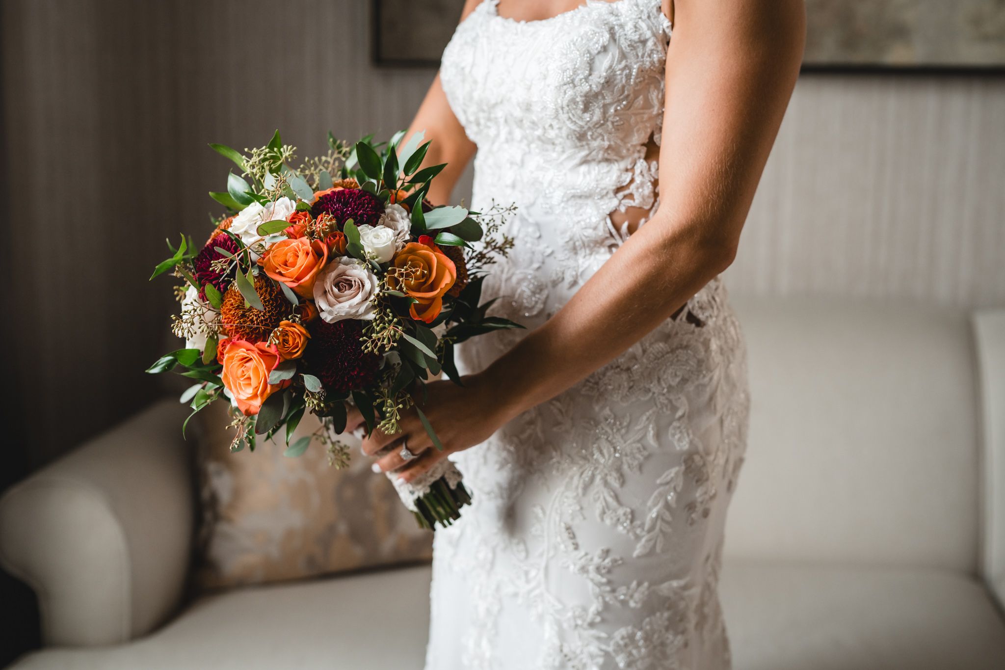 Close-up details of white lace wedding dress and vibrant orange and burgundy bridal bouquet.