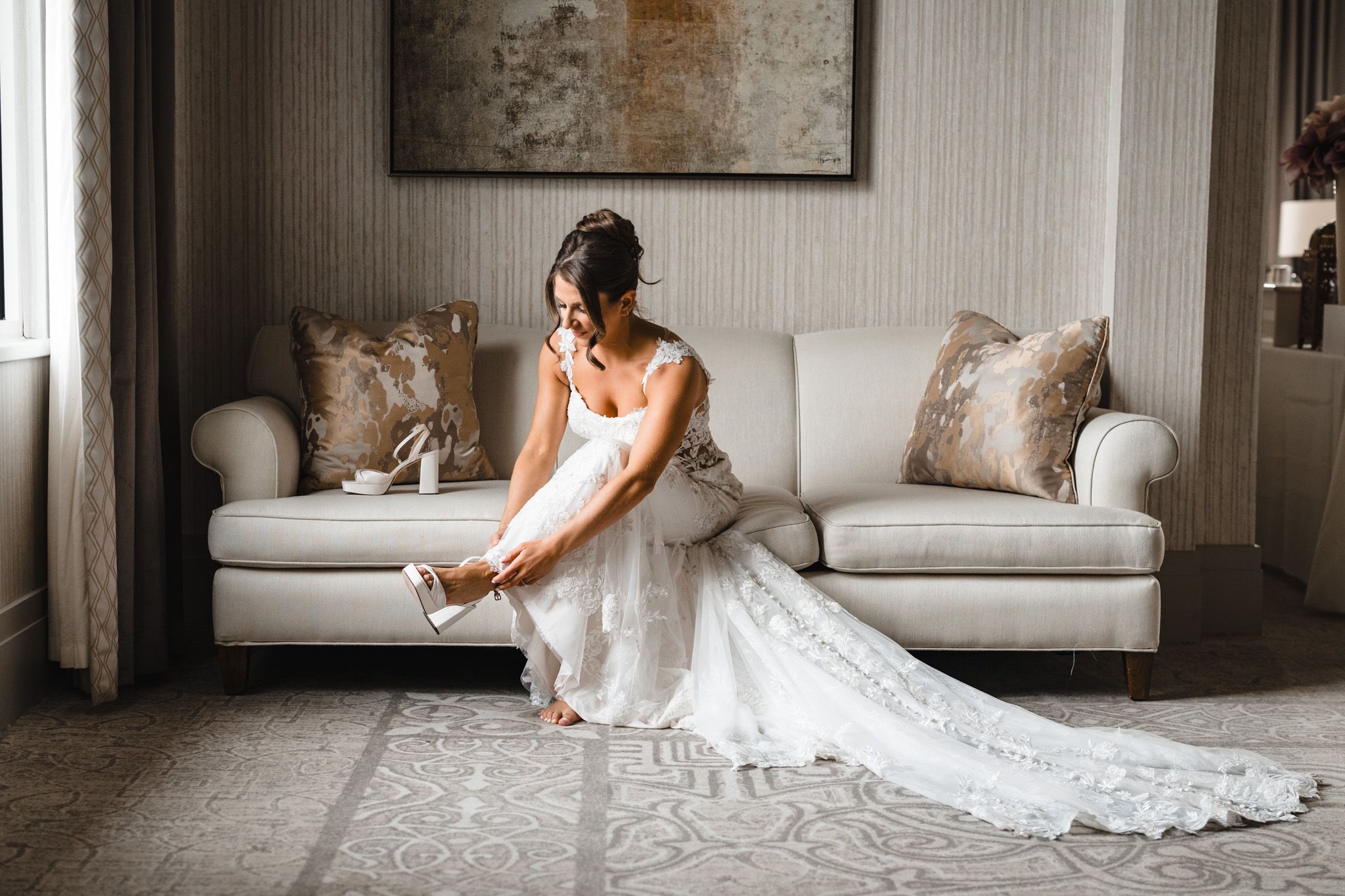 Wedding dress with intricate lace details spreads across white sofa in natural window light.