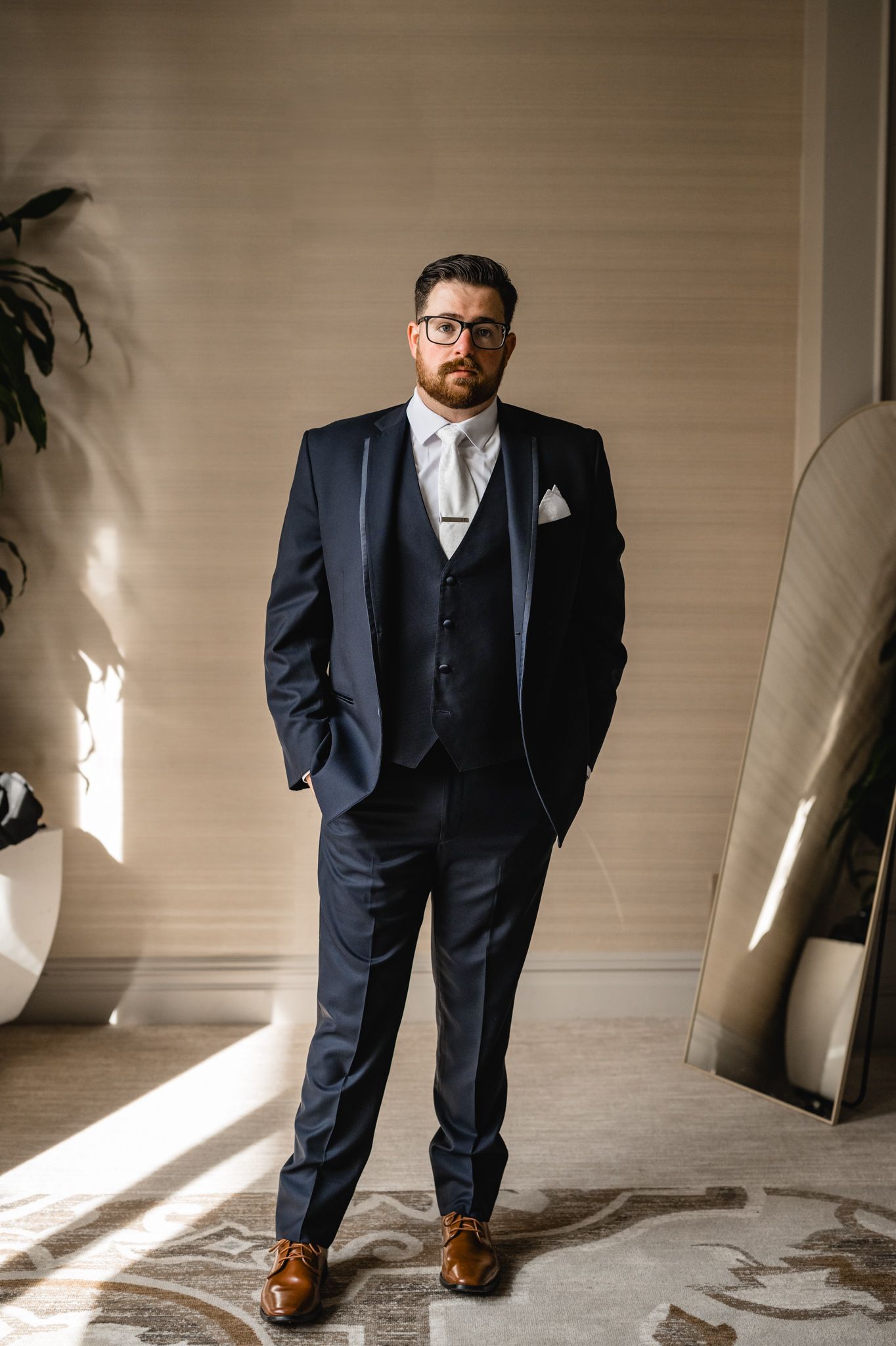 A groom in a navy three-piece suit and brown shoes stands in dramatic window lighting.