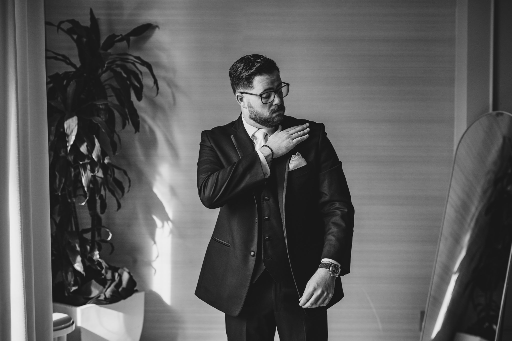 Artistic black and white portrait of a groom in formal attire standing by a window.