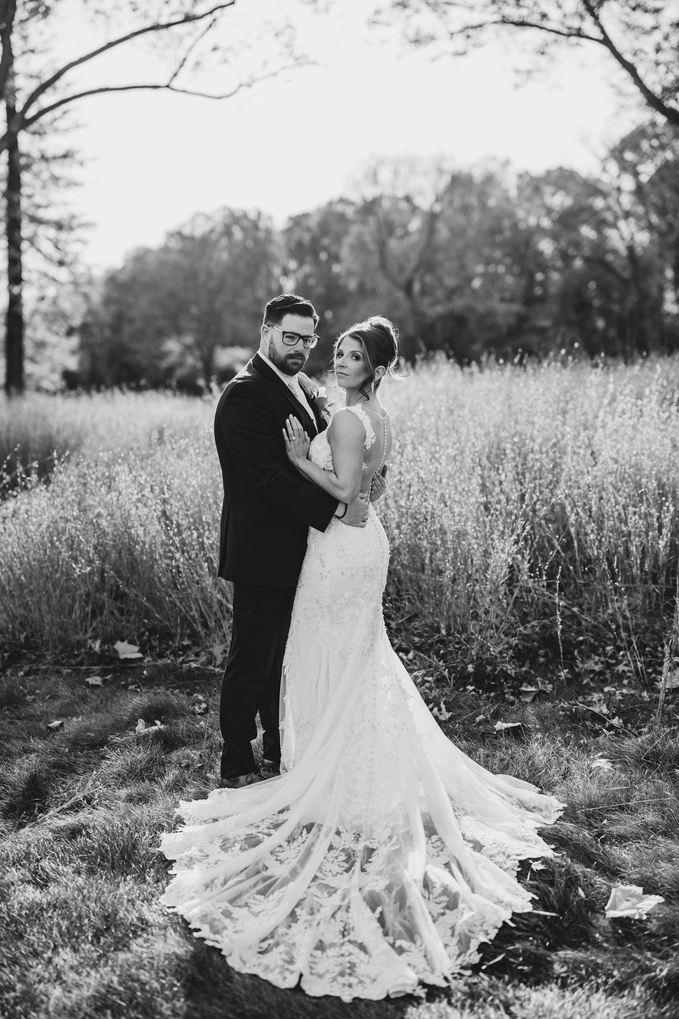 Black and white wedding portrait showing bride's lace dress train in tall grass field.