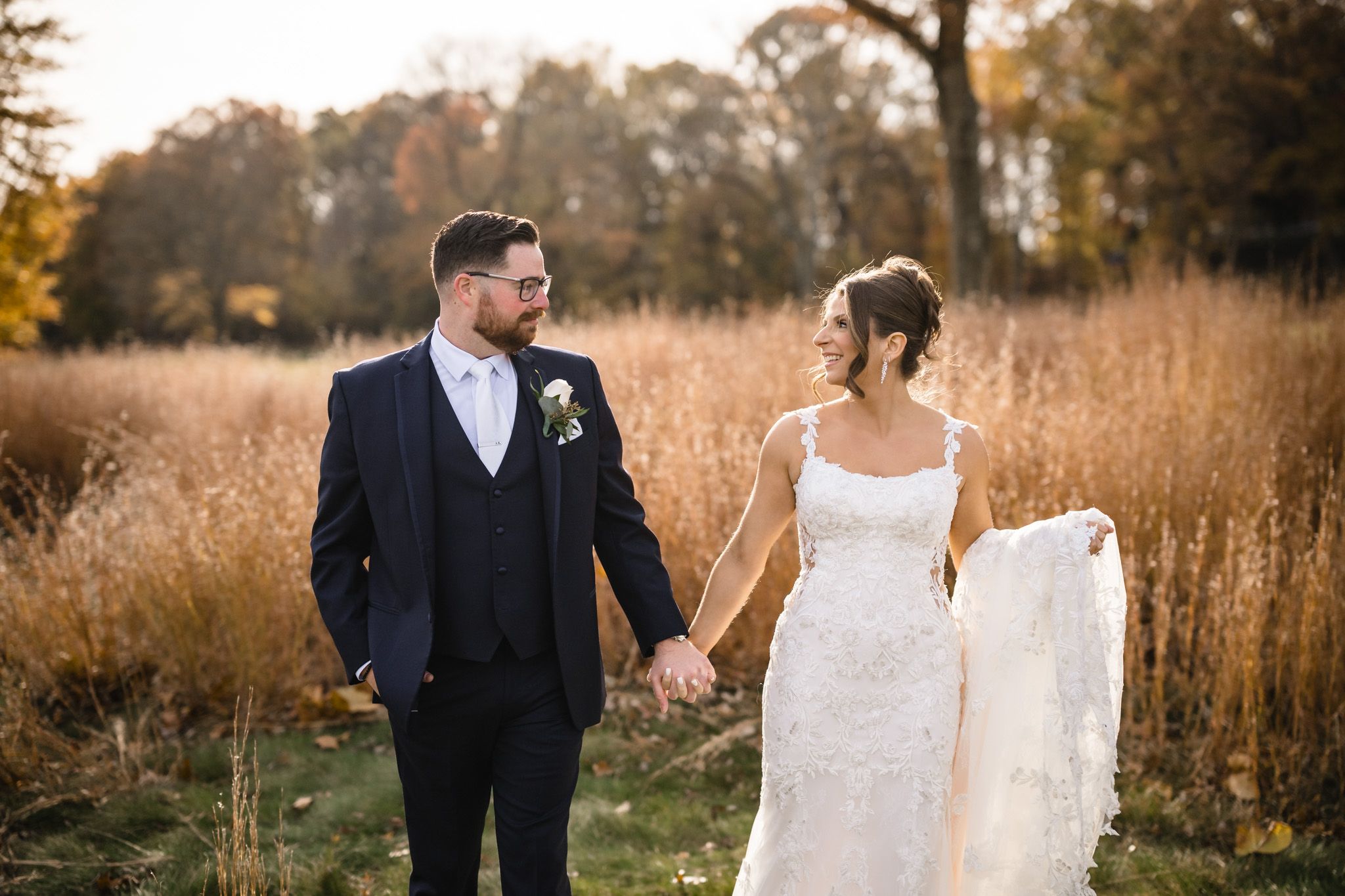 Bride in lace gown and groom in dark suit walk through fall meadow.