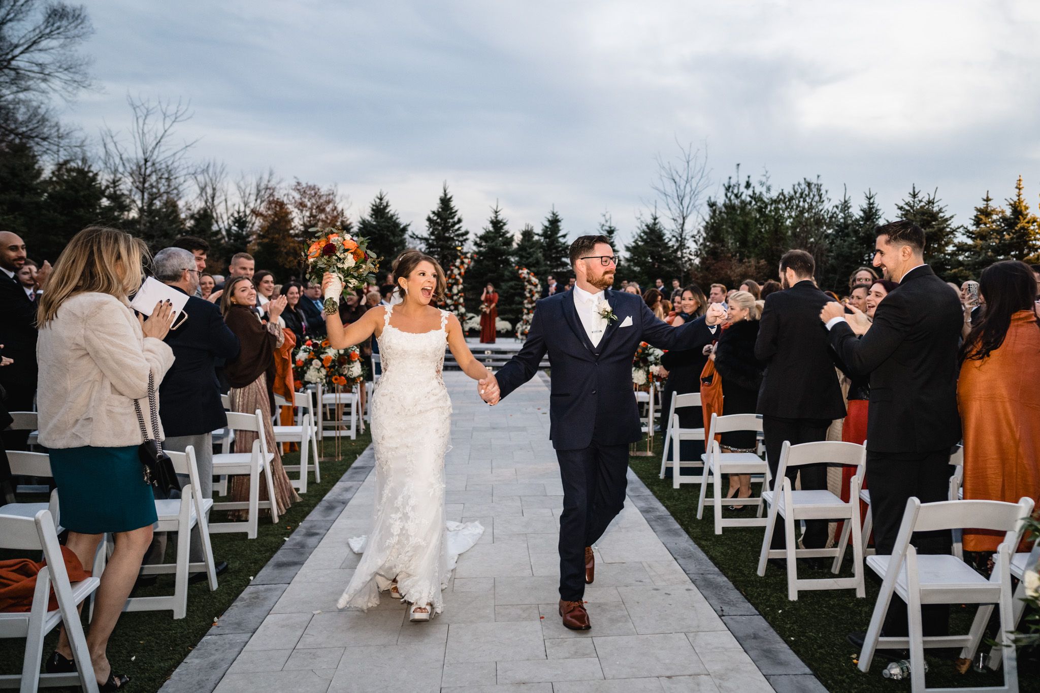 Joyful recessional along outdoor wedding aisle with white chairs and autumn decor.