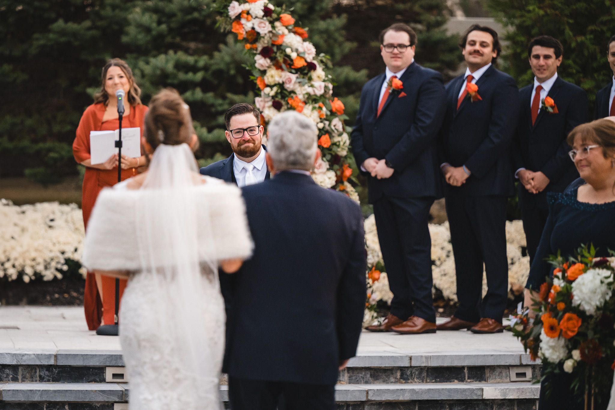 Outdoor wedding ceremony with orange and white floral arrangements and evergreen backdrop.