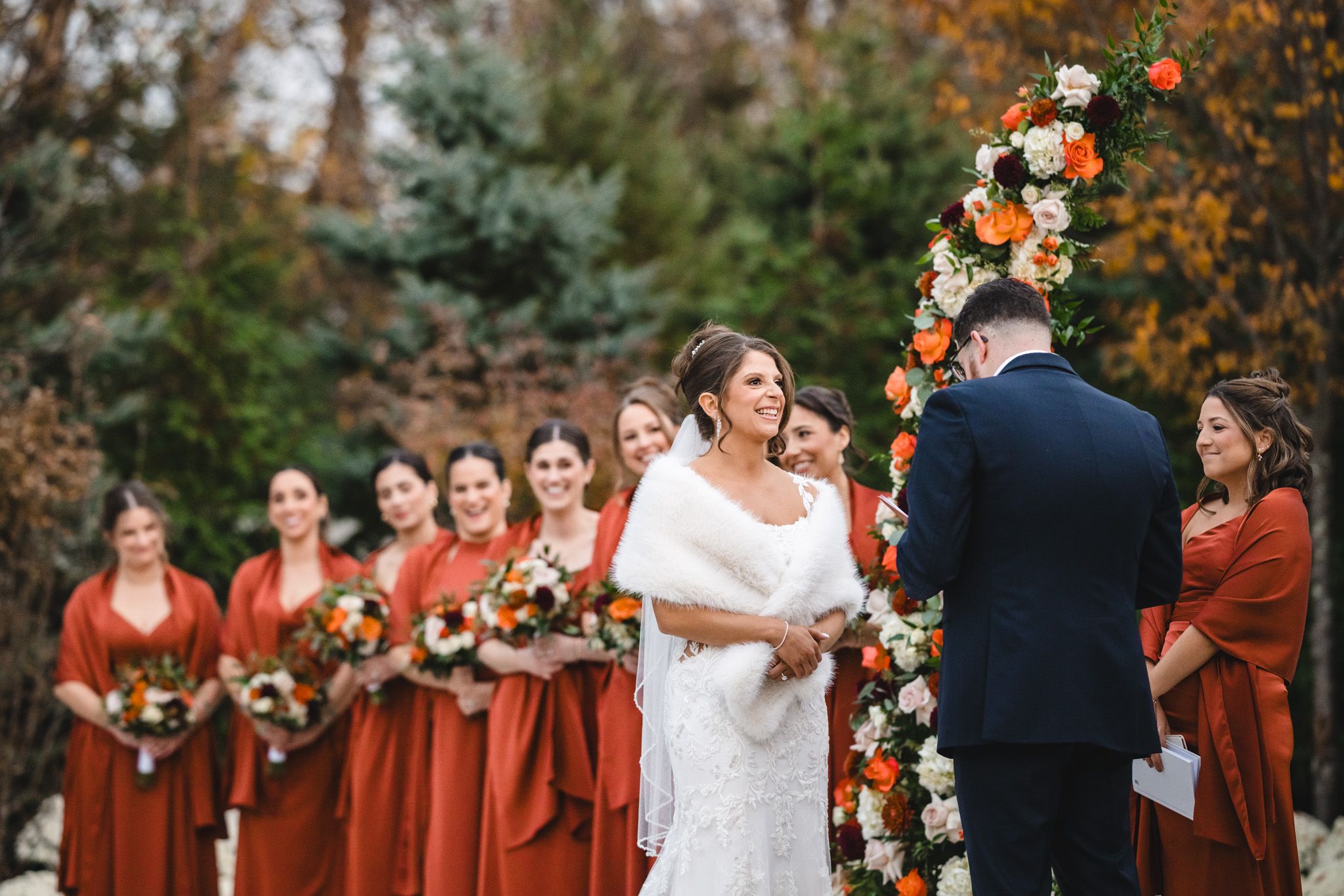 Wedding ceremony outdoors with autumn colors and floral arch decorations in burnt orange and white.