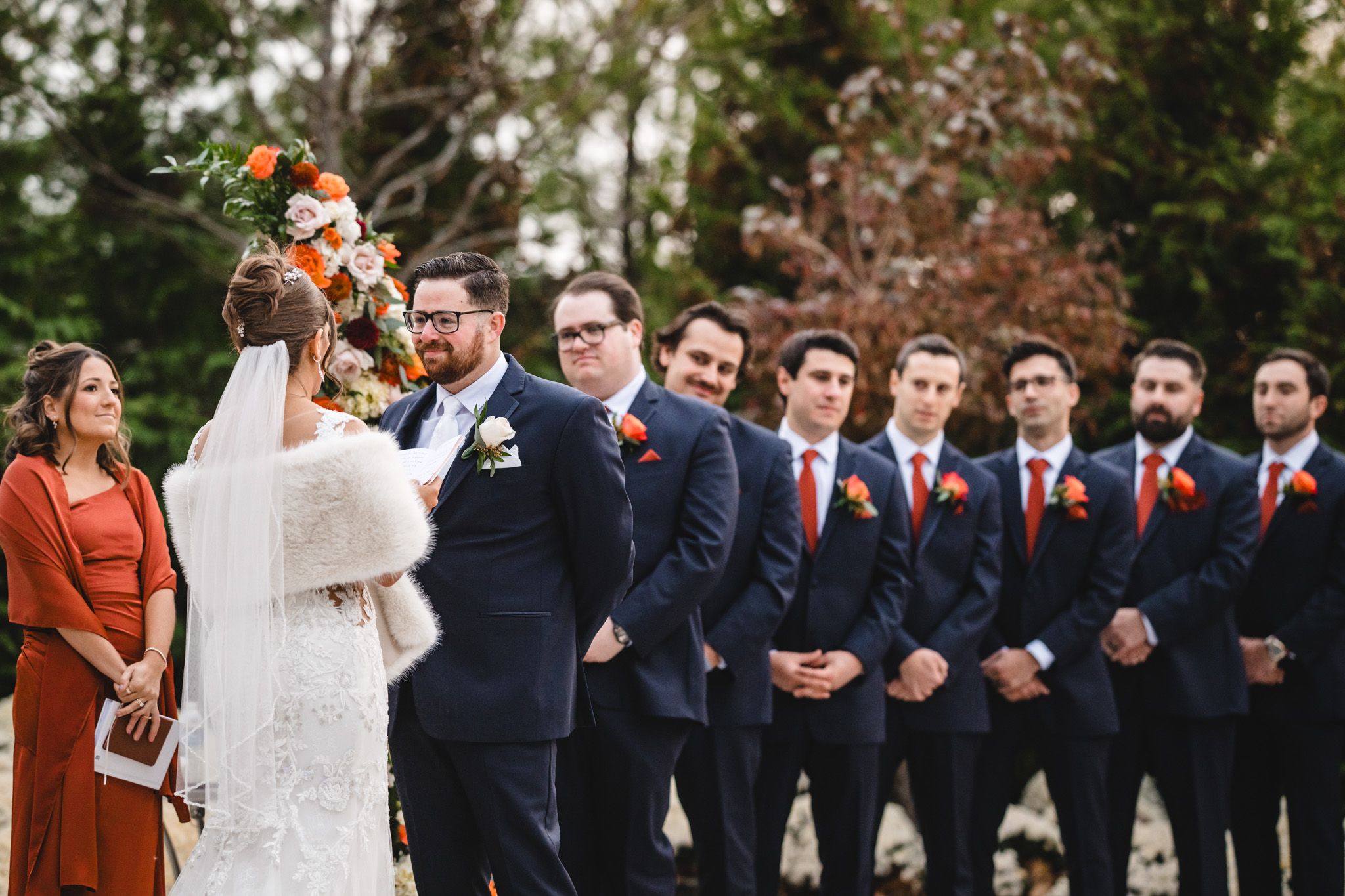 Wedding party lined up outdoors with bridesmaids in rust-colored dresses and groomsmen in navy suits.