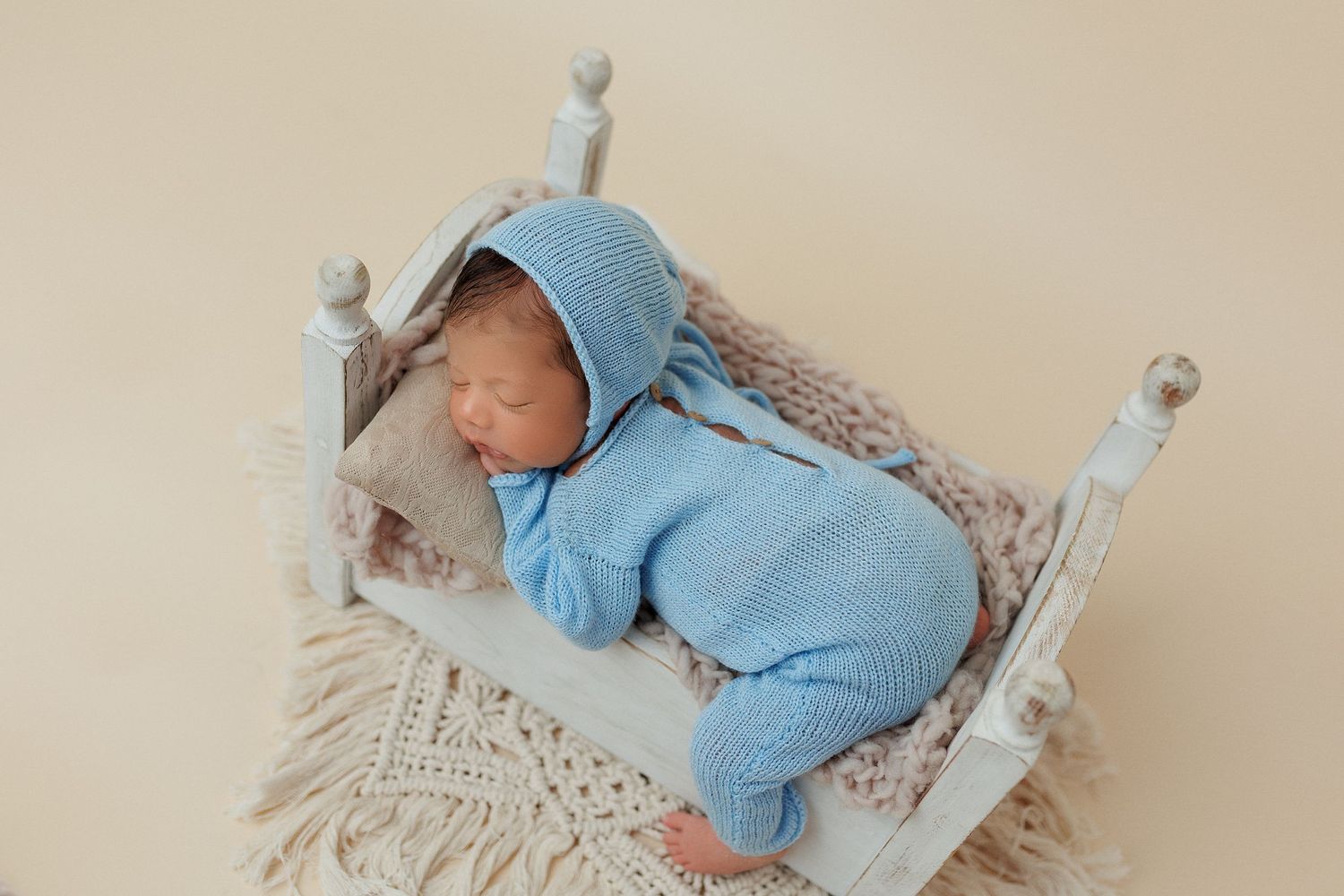 Sleeping newborn in blue outfit on white miniature bed with knitted blanket.