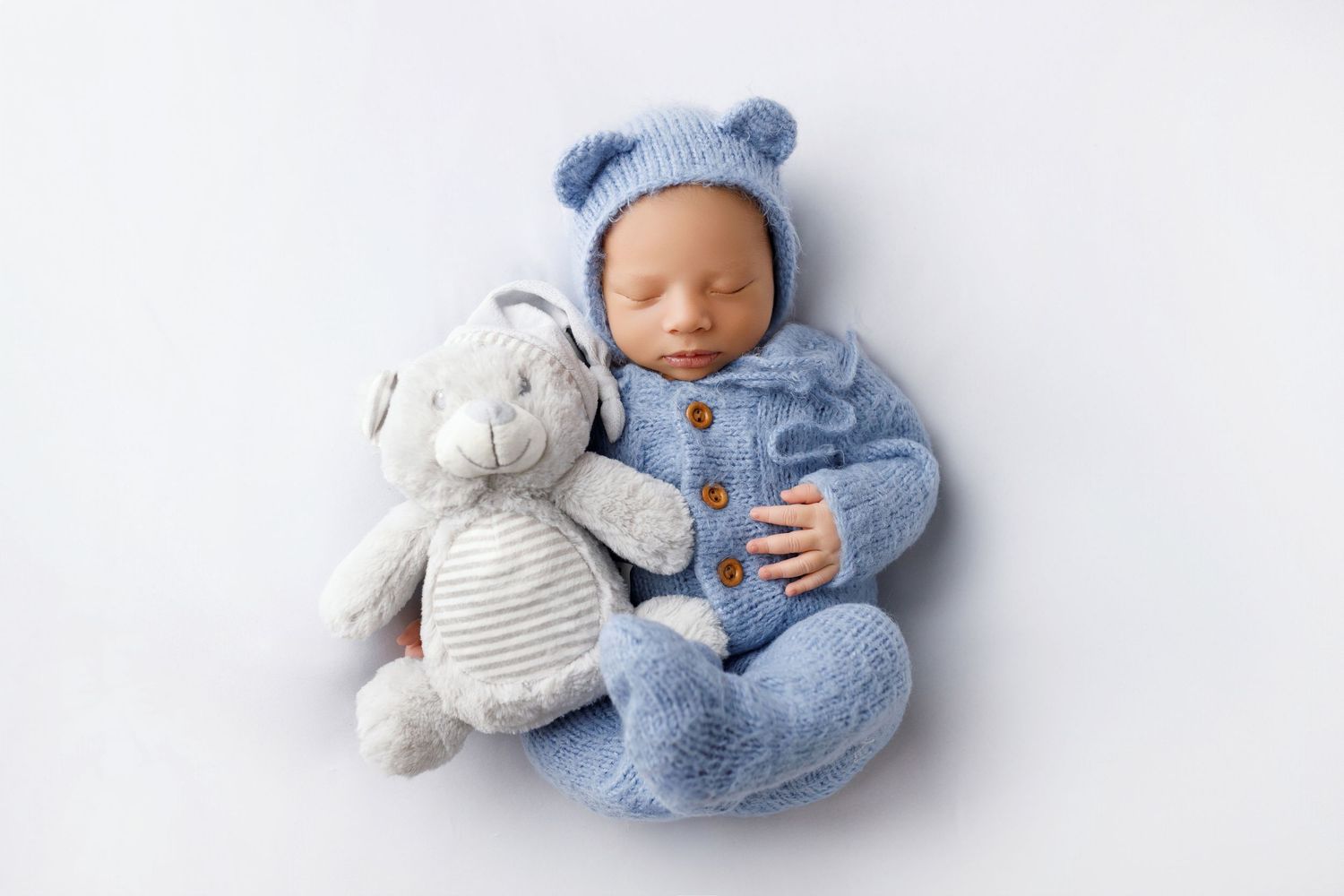 Sleeping newborn in blue knit outfit curled up with plush elephant toy on white backdrop.