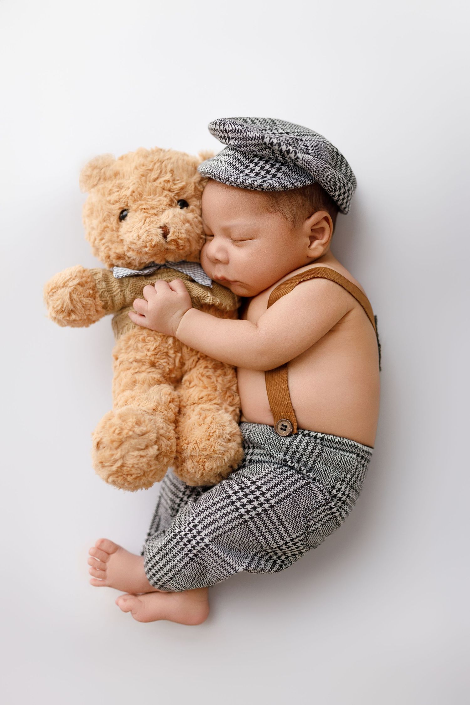 A sleeping baby in plaid pants and flat cap snuggles with a tan teddy bear on a white background.
