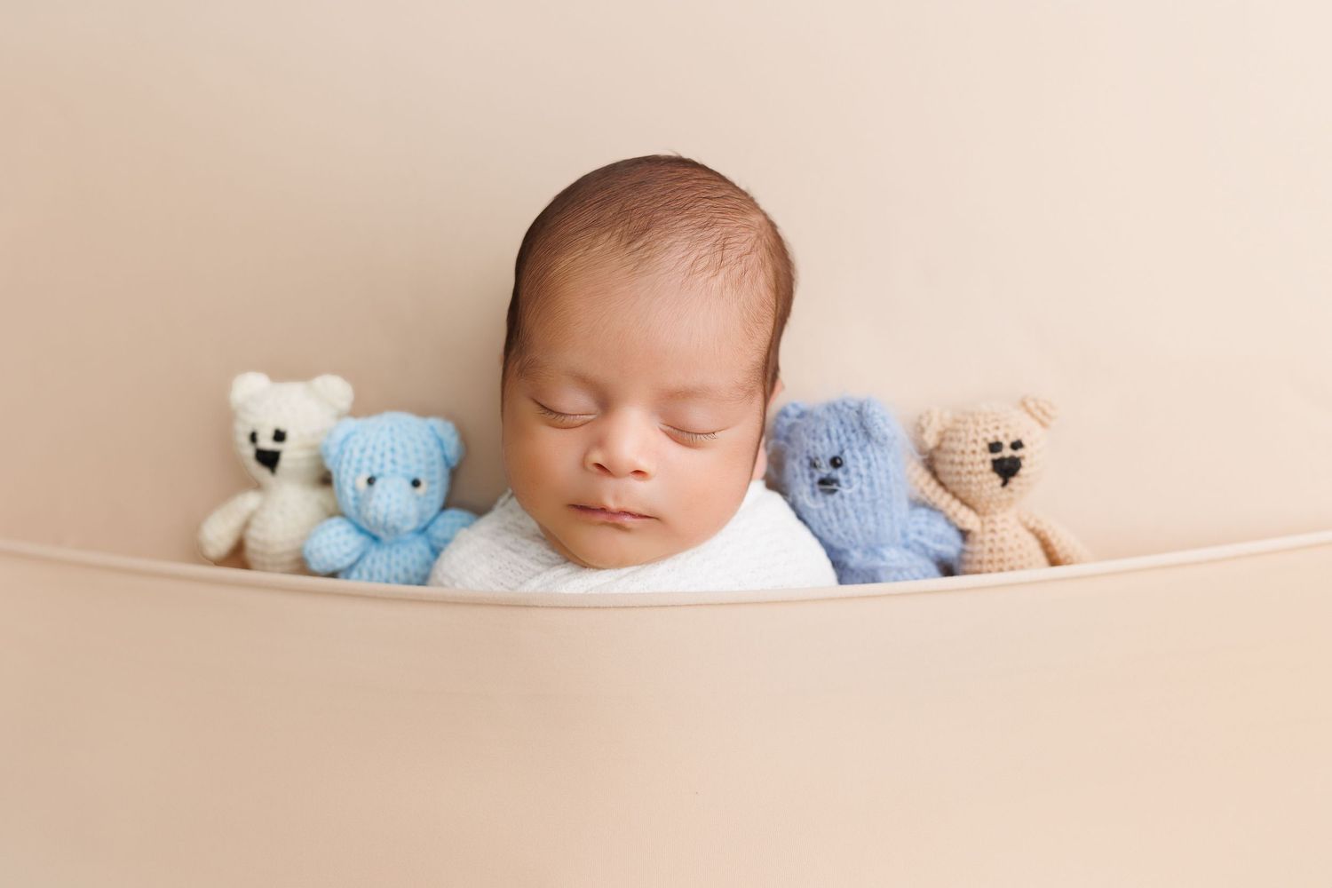 A sleeping newborn baby lies peacefully on cream-colored fabric surrounded by small blue and white teddy bears.