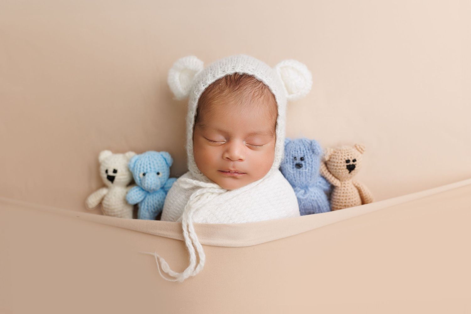 Sleeping newborn baby wearing white hat lies peacefully with soft teddy bears and plush toys in neutral tones.