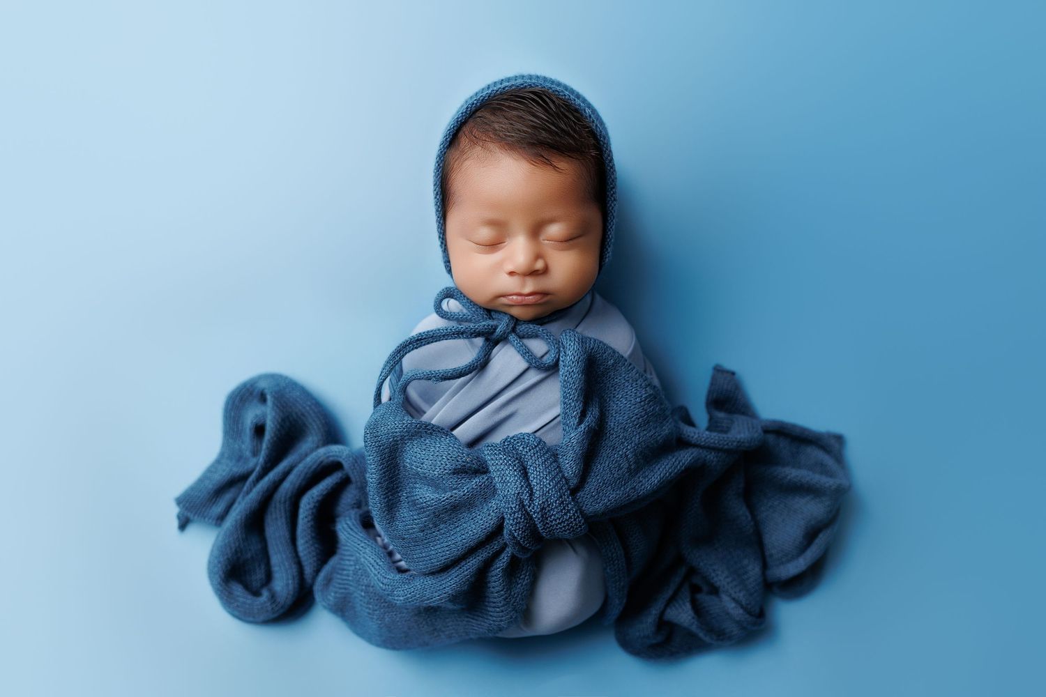 Newborn baby in blue swaddle cloth sleeping peacefully against light blue background.