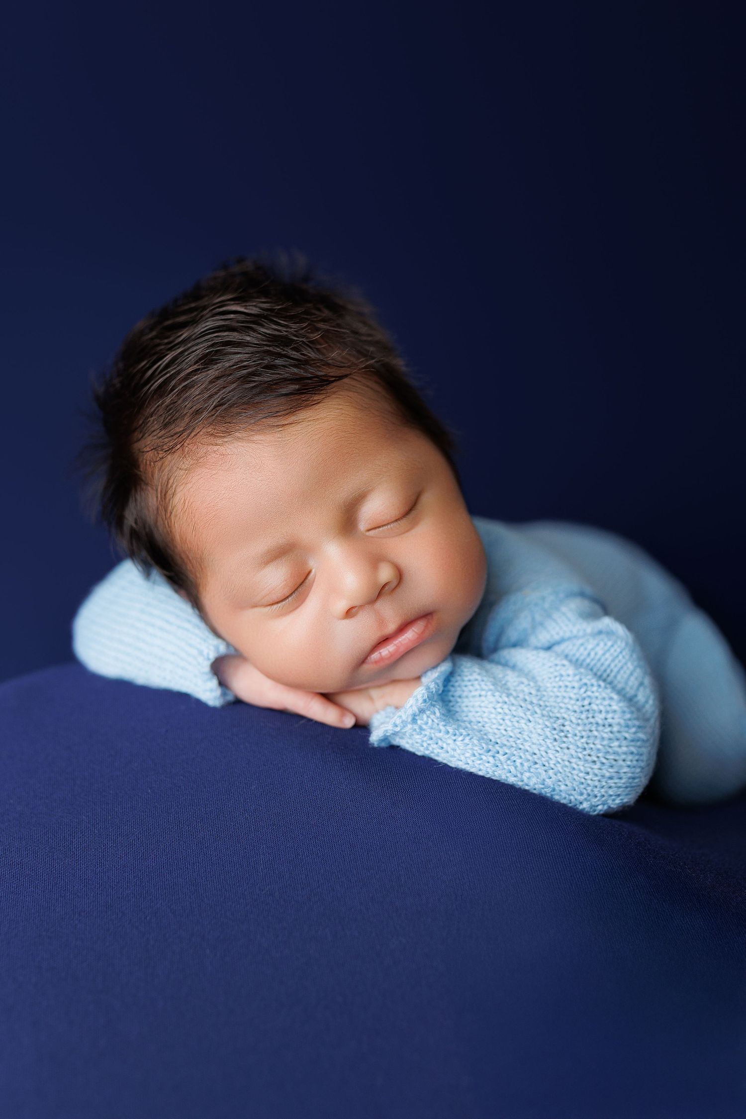 Peaceful sleeping newborn baby wrapped in soft blue blanket against dark navy background in professional portrait.