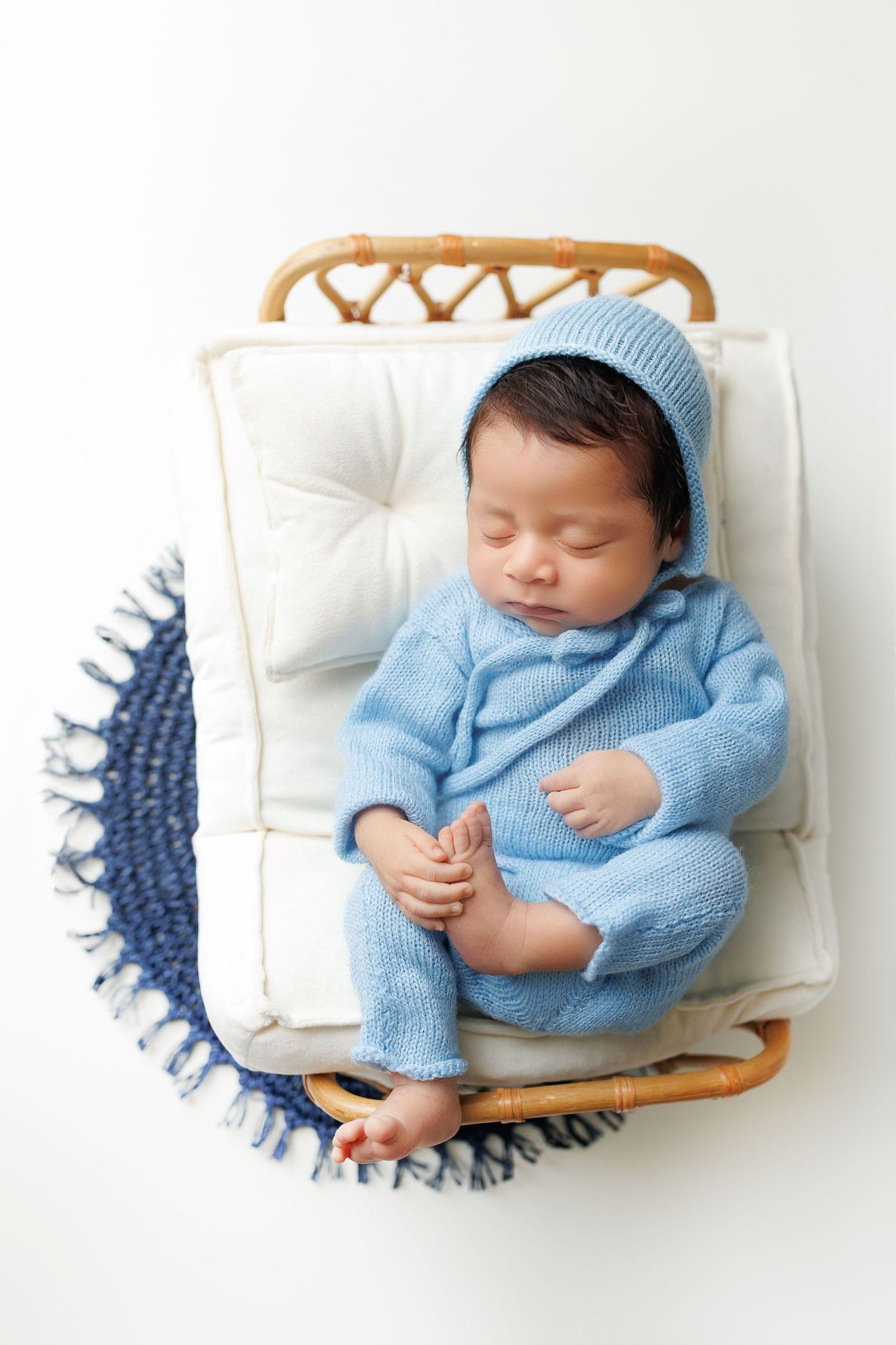 A sleeping baby wearing a light blue knit outfit lies peacefully on a white cushion in a woven basket.