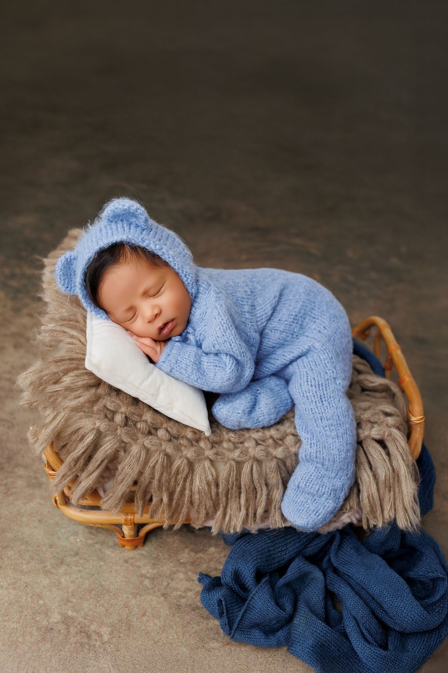A sleeping newborn wearing a soft blue sweater rests peacefully on a textured brown blanket with navy blue fabric underneath.