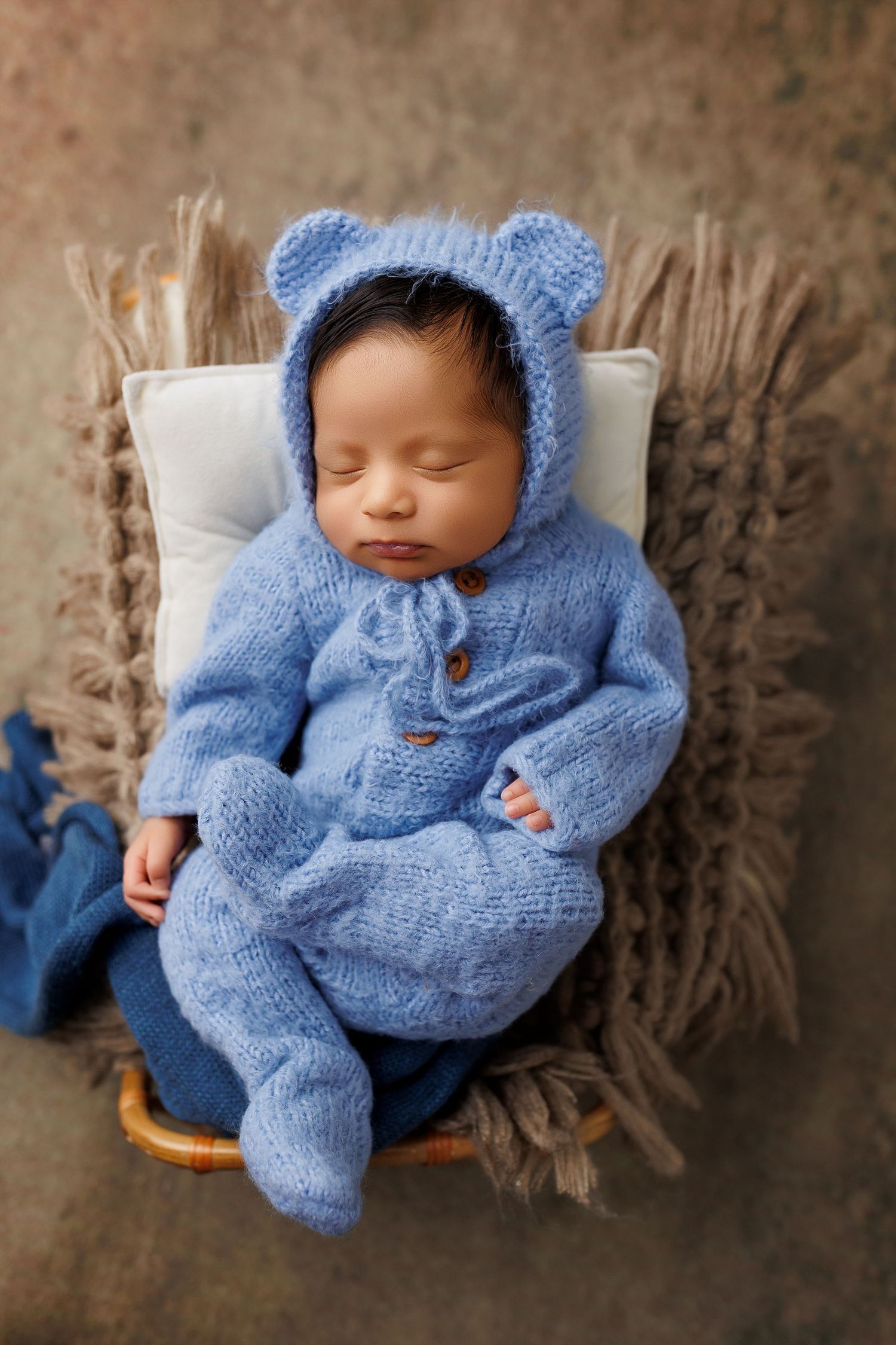 Sleeping baby wearing cozy blue knitted bear outfit lying on textured brown blanket in newborn photography session.