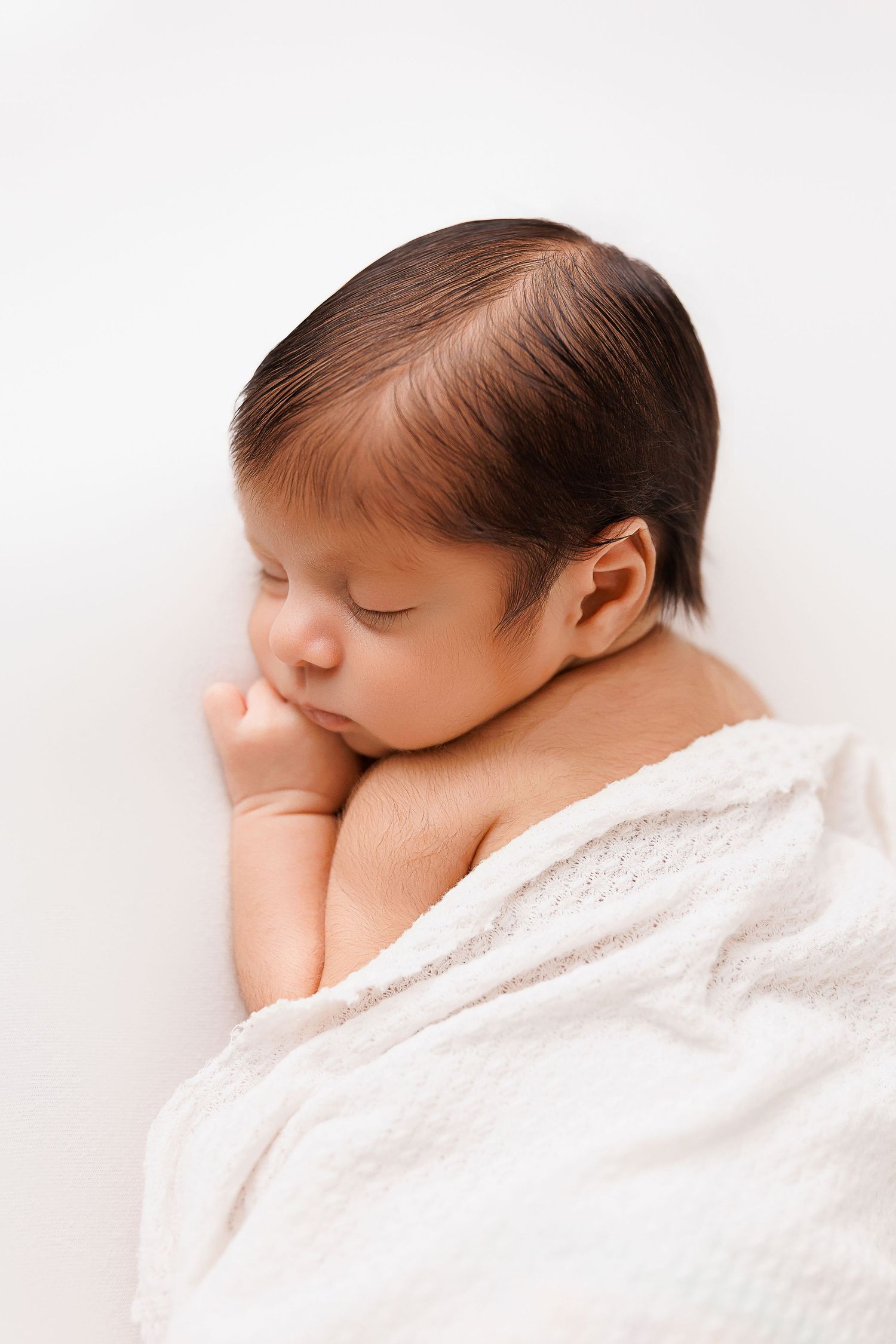 Sleeping newborn baby wrapped in soft white blanket resting peacefully against light background.