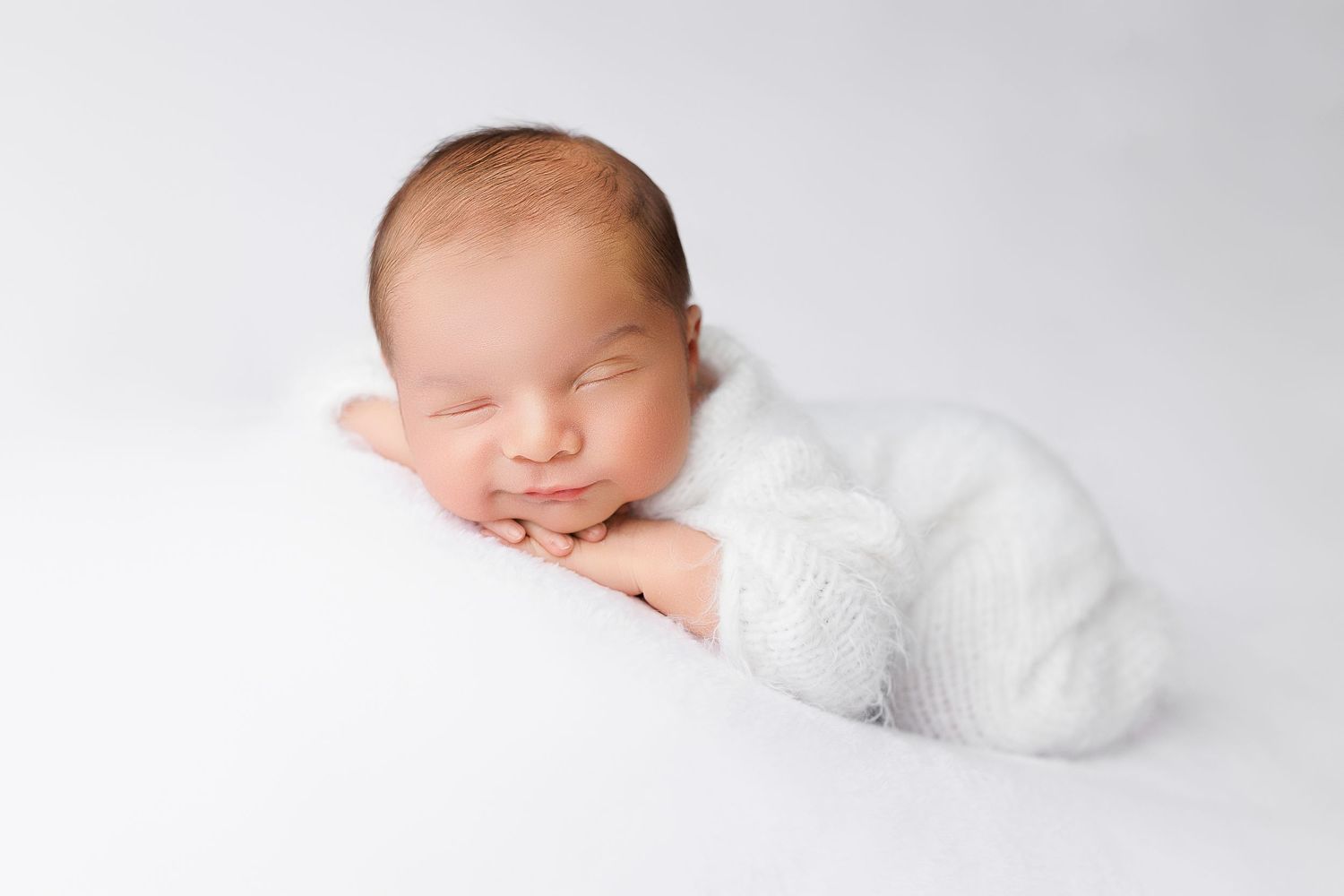 A peaceful sleeping newborn wearing white lies on a soft white backdrop in a professional studio setting.