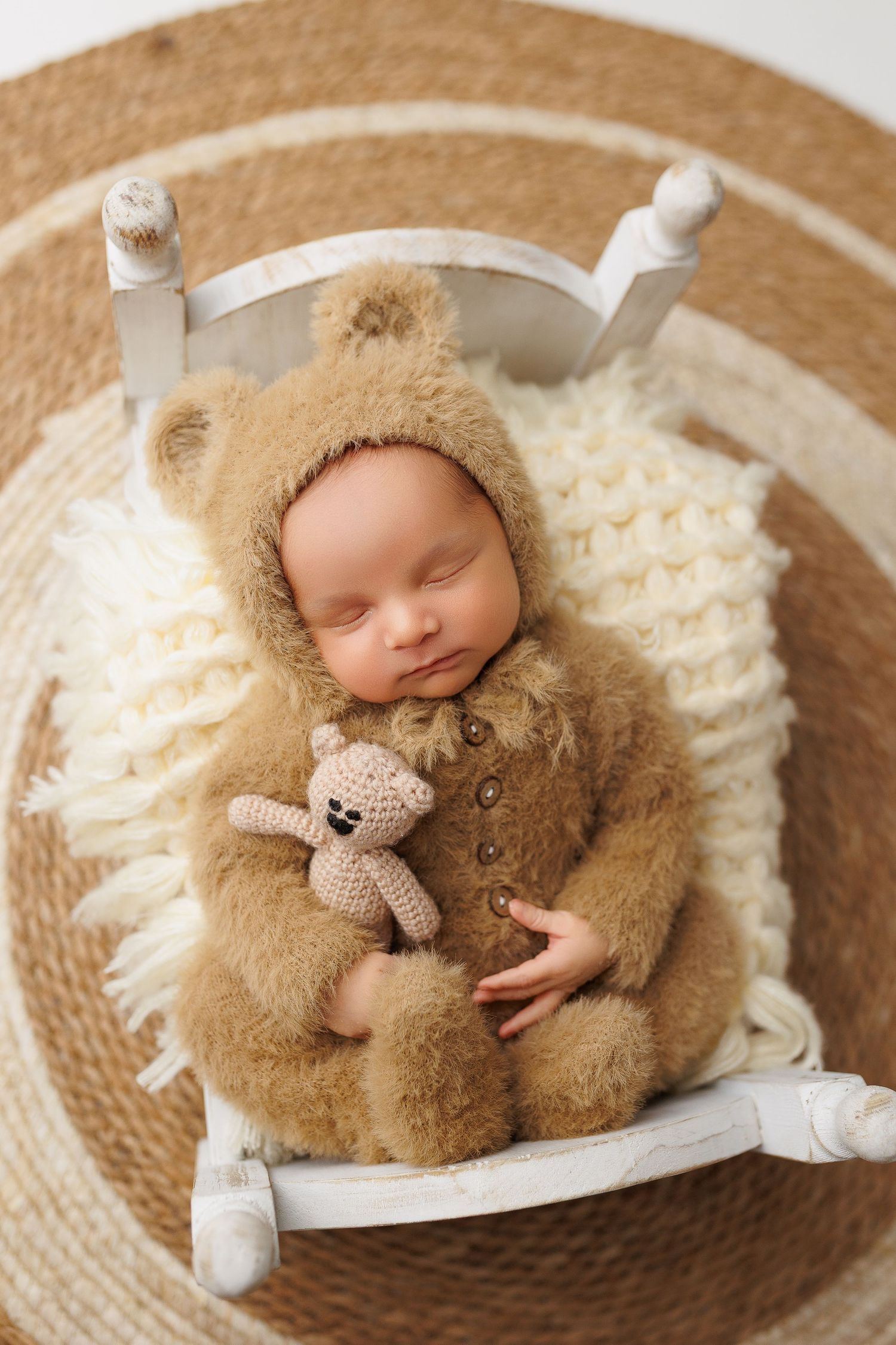 Sleeping newborn in cozy brown bear outfit holding tiny teddy bear on white chair with woven rug background.