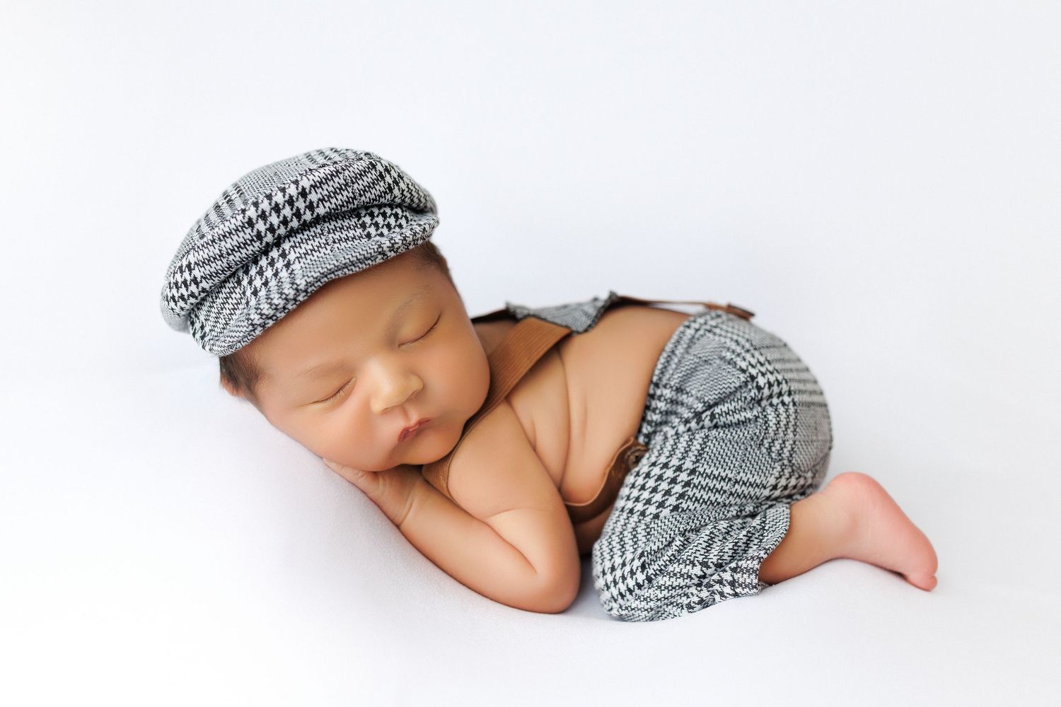 A sleeping newborn baby wearing a plaid newsboy cap rests peacefully on white bedding.