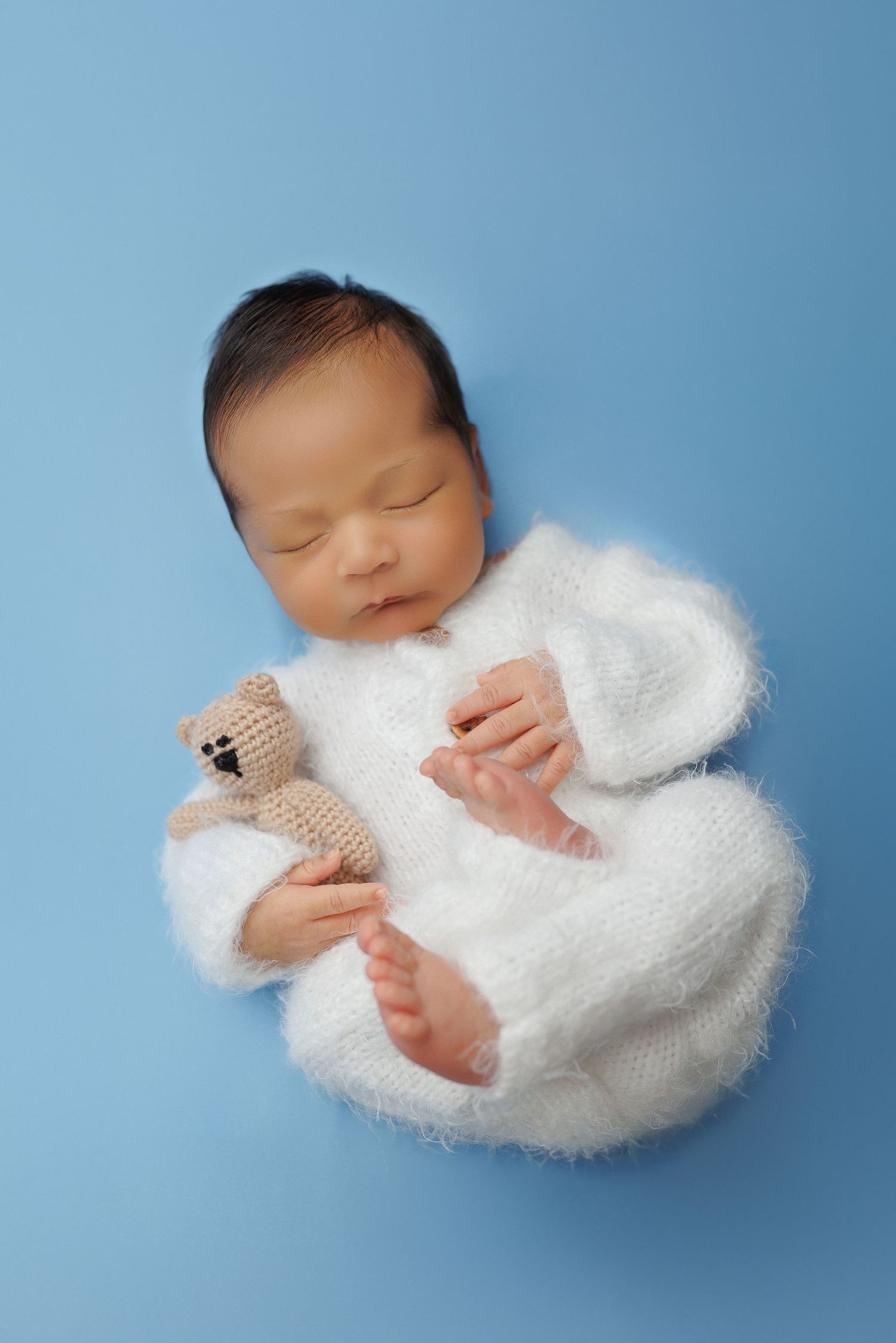 A sleeping newborn wrapped in soft white fabric holds a tiny teddy bear while lying on a blue background.