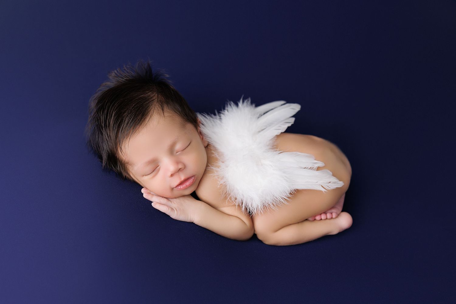 A sleeping baby wearing angel wings lies peacefully on a dark blue backdrop in a series of artistic newborn portraits.