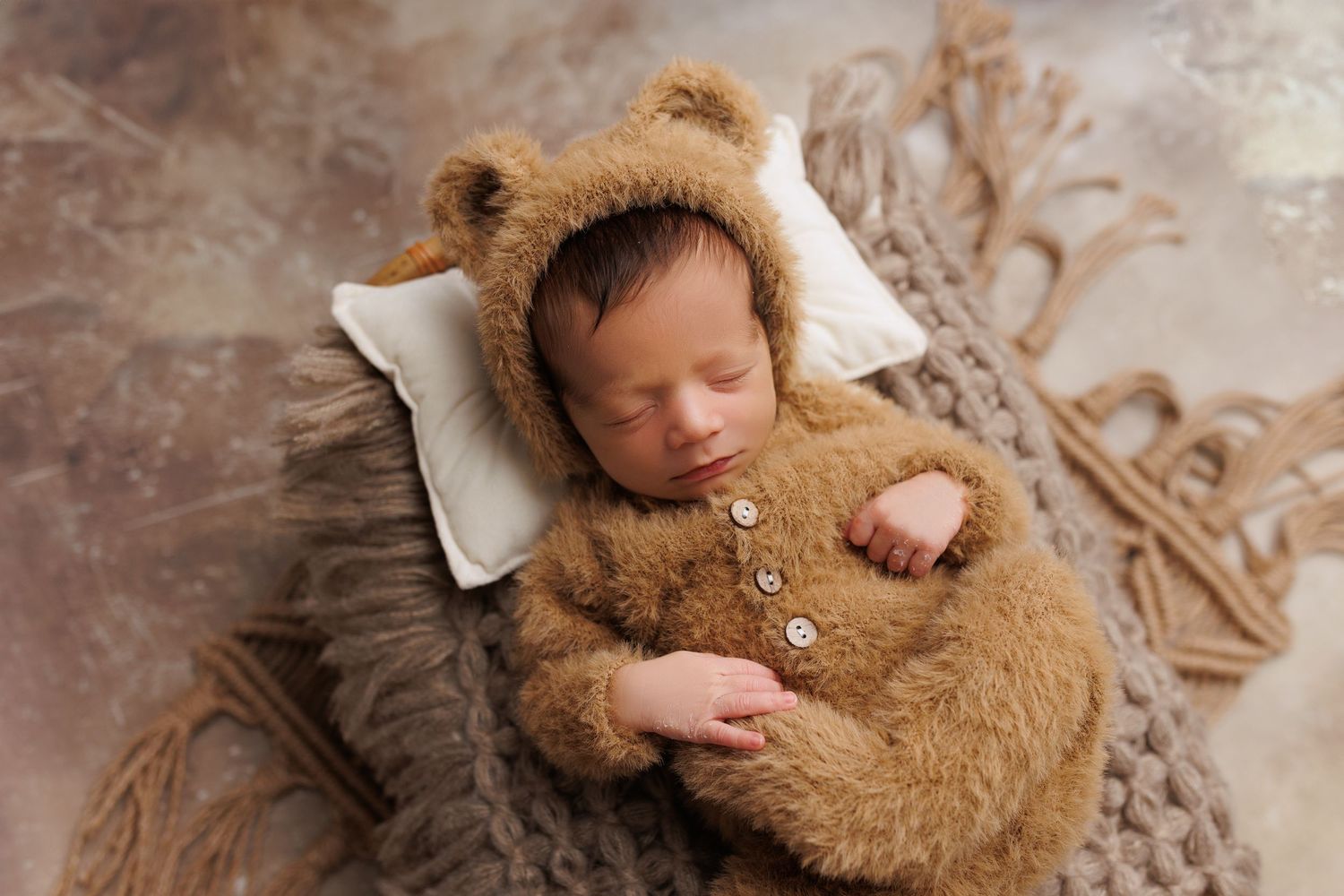 Newborn baby sleeping peacefully while cuddling a soft brown teddy bear on textured beige backdrop.