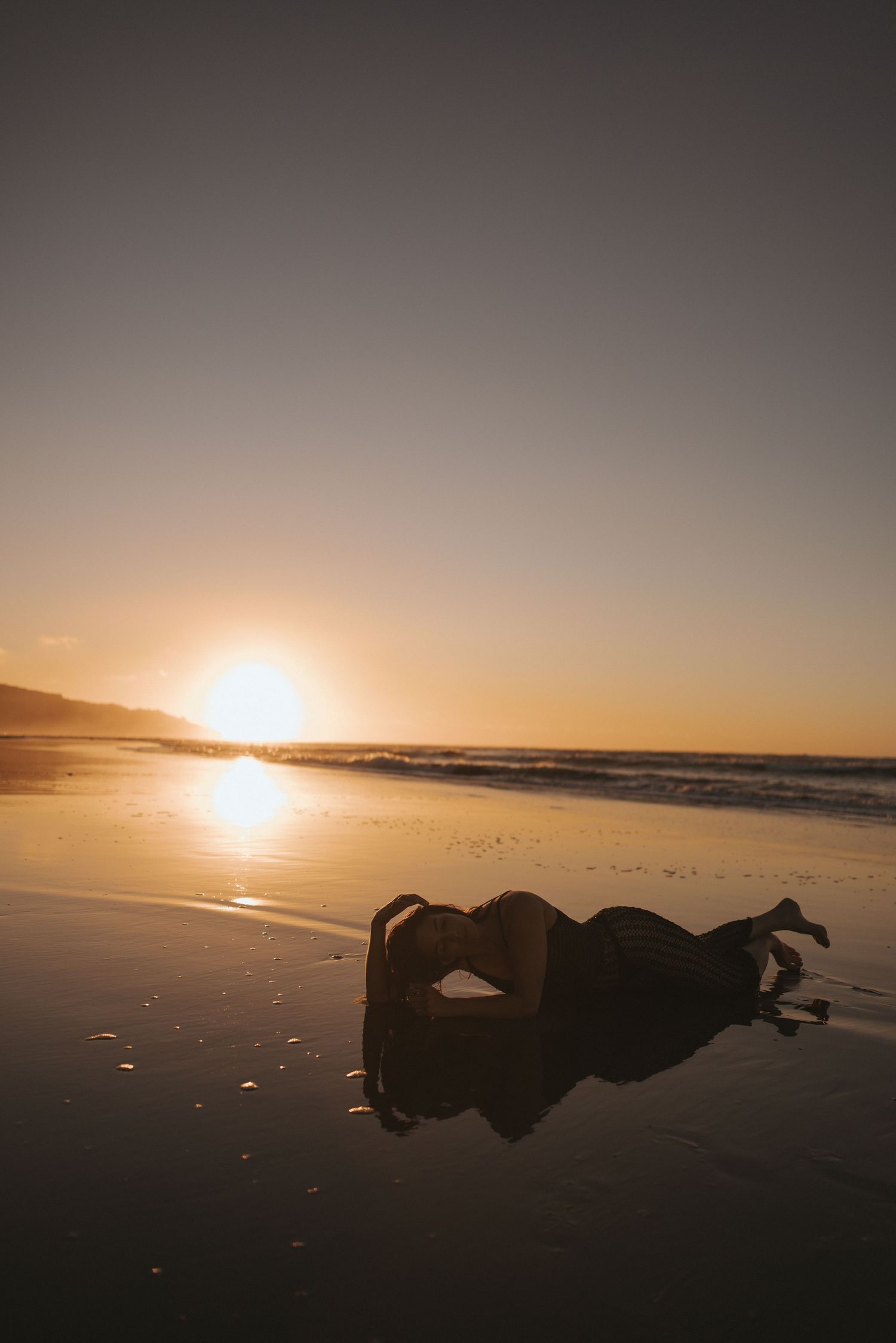 Silhouette of a person lying on a wet beach at golden sunset with ocean waves in the background in New Zealand.