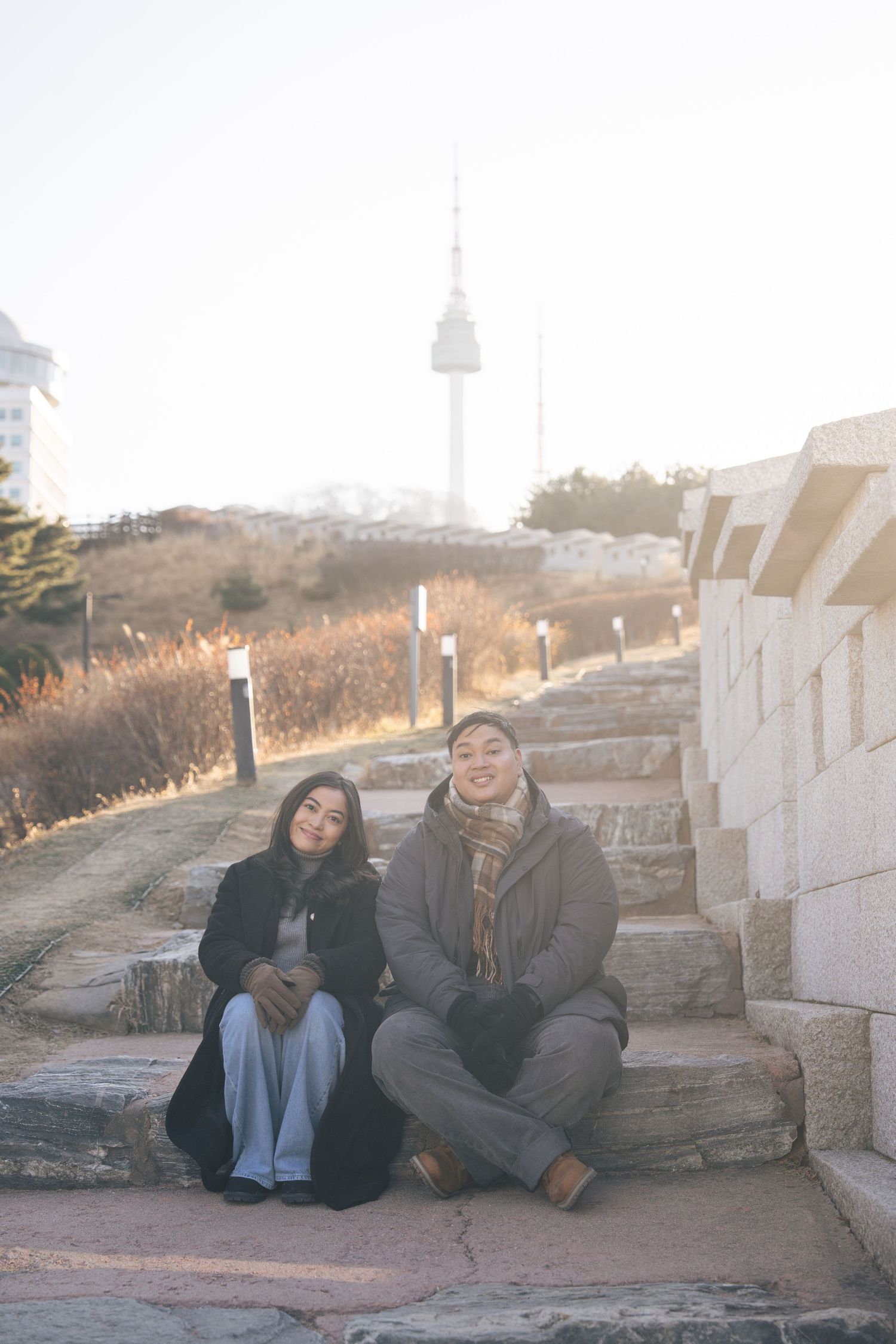 Two people in warm winter clothing sitting together on stone steps at sunset in an outdoor setting.