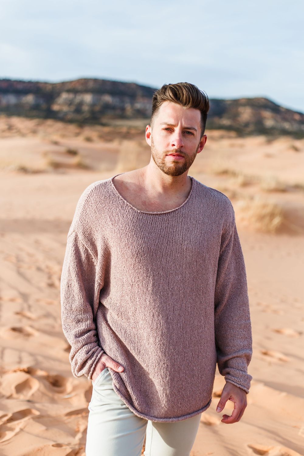 A person in a mauve sweater poses against desert landscape with mountains in the background.