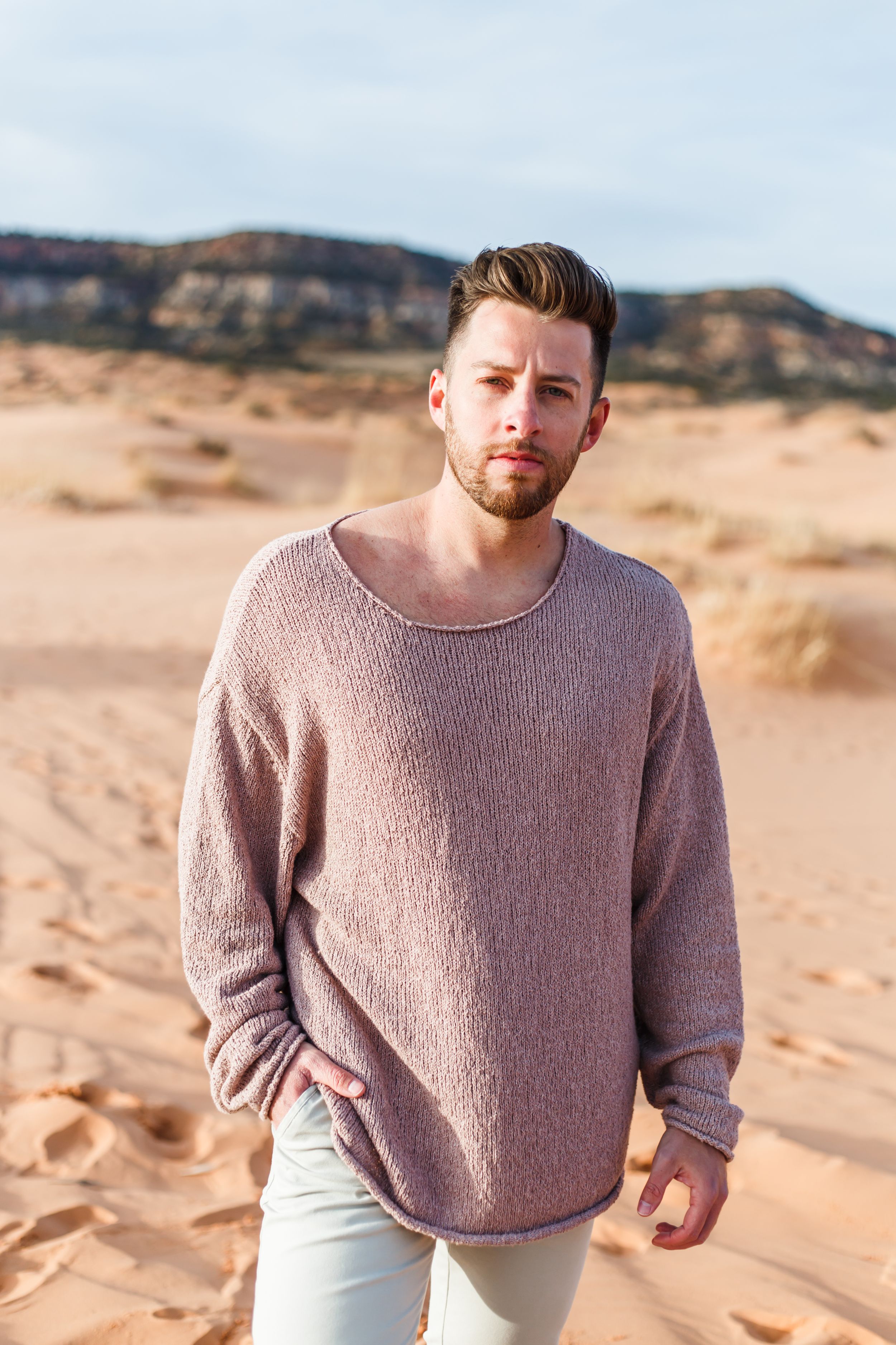 A person in a mauve sweater poses against desert landscape with mountains in the background.