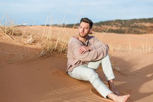 Person in light colored clothing sits on sand dunes during golden hour at a desert landscape.