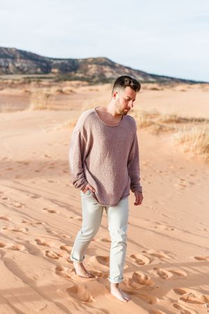Person wearing a mauve sweater and light pants walking on sandy desert dunes with mountains in background.