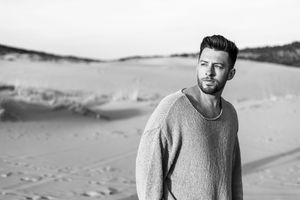 A person wearing a light colored sweater stands against a desert landscape background with rolling sand dunes.