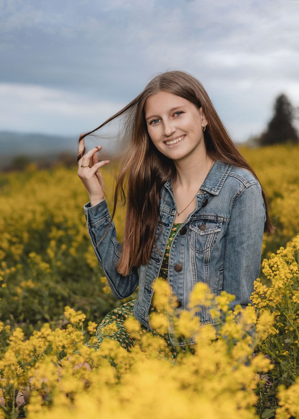Woman in denim jacket poses in field of bright yellow wildflowers on cloudy day.