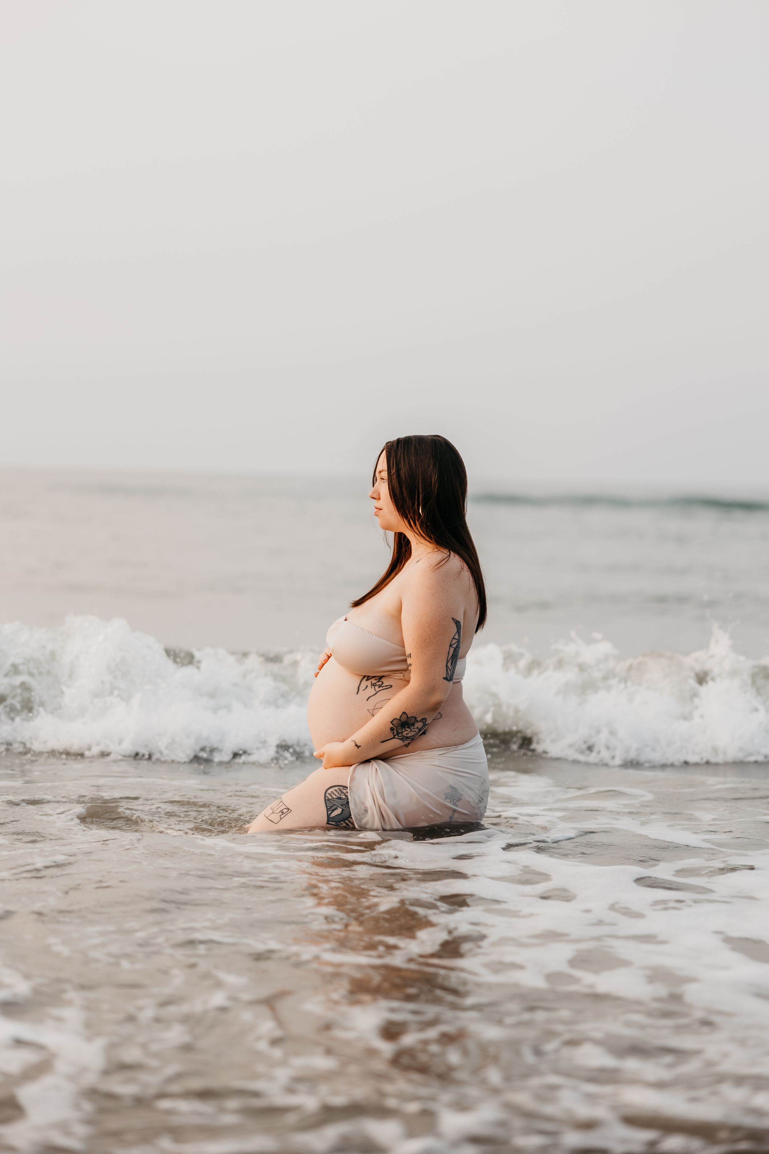 An expectant mother in a flowing dress sits peacefully in the ocean waves during sunset.