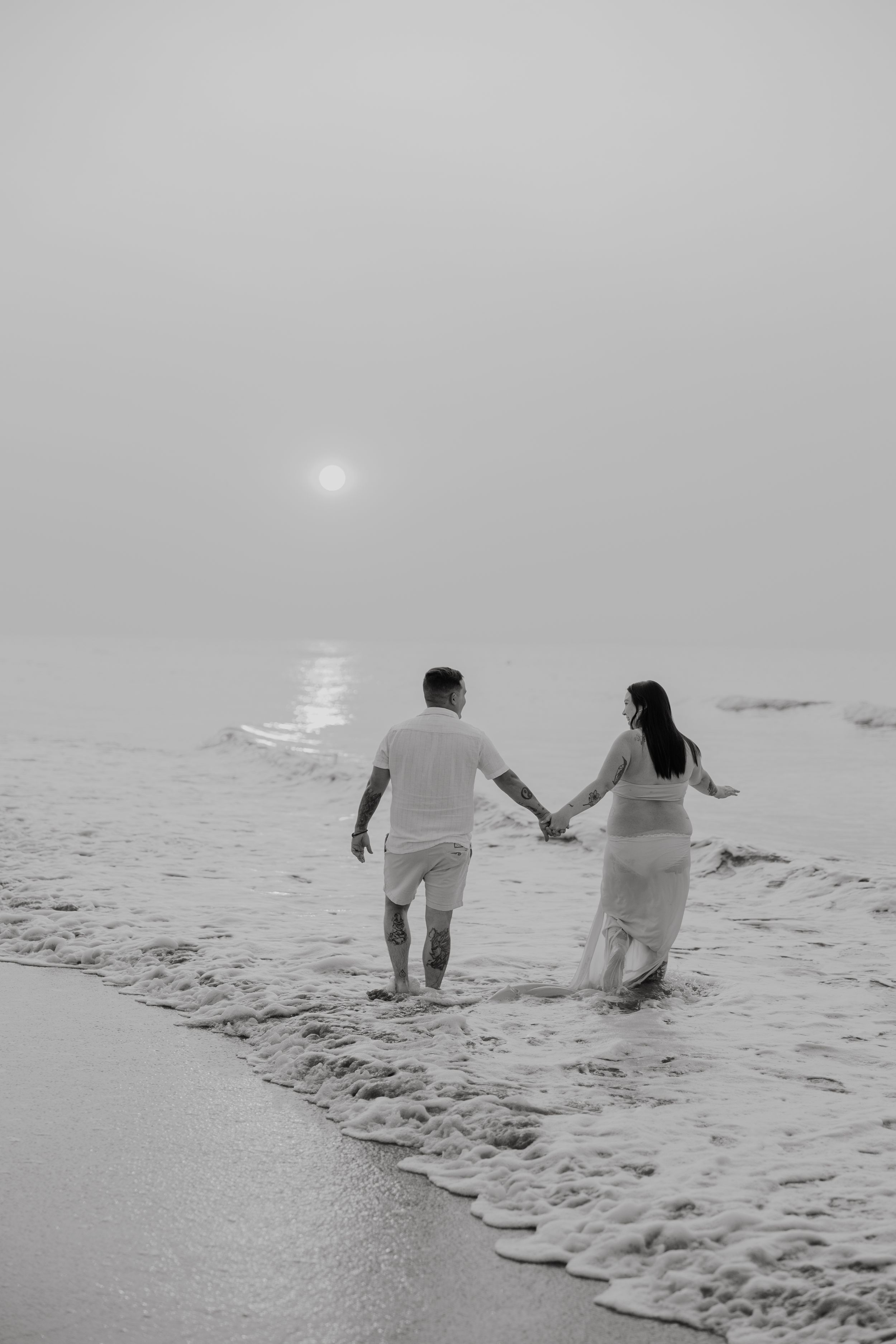 A couple holds hands while walking through ocean waves on a misty beach.