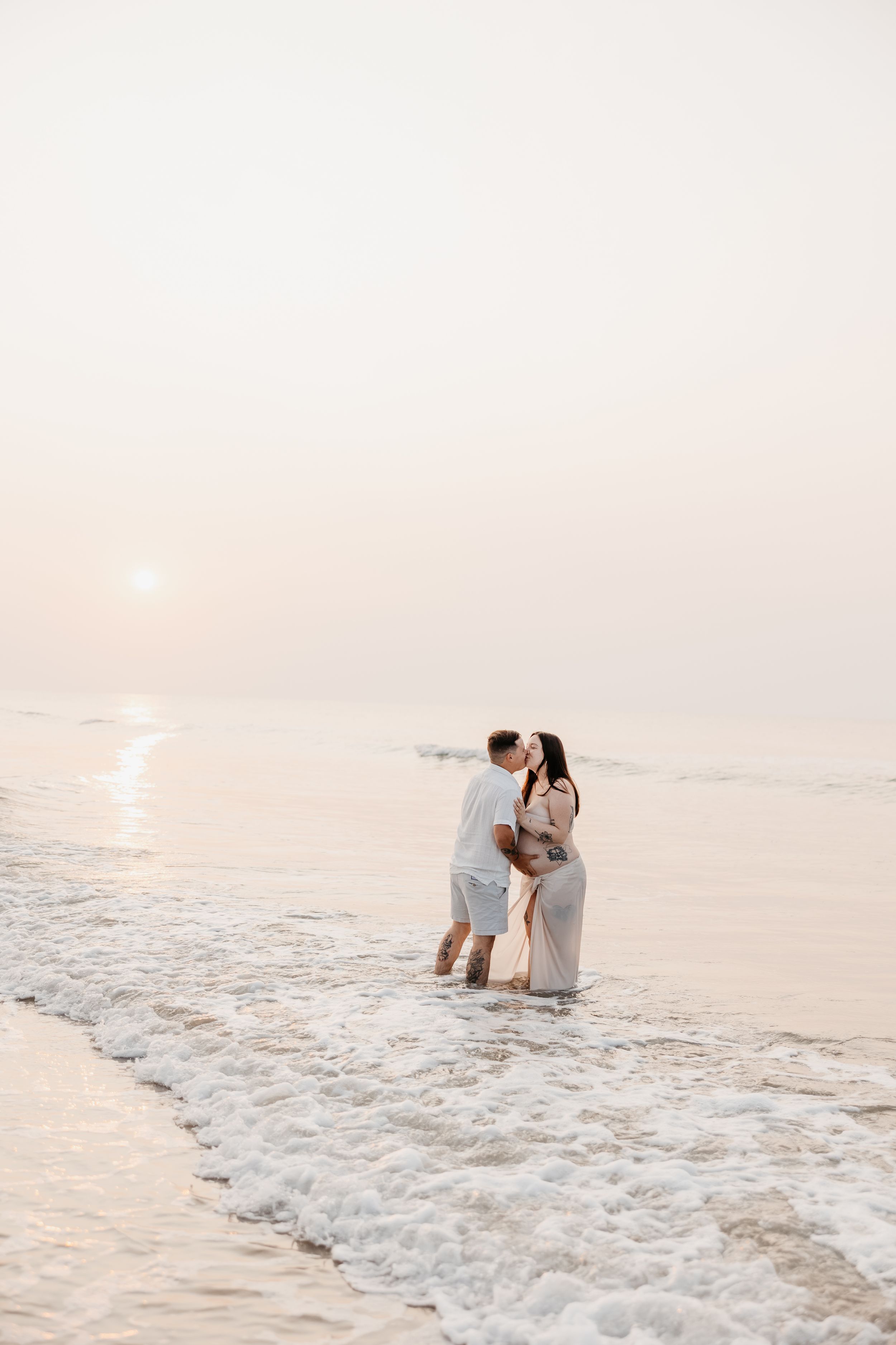 A romantic couple embraces in the shallow ocean waves during a sunset beach photoshoot.