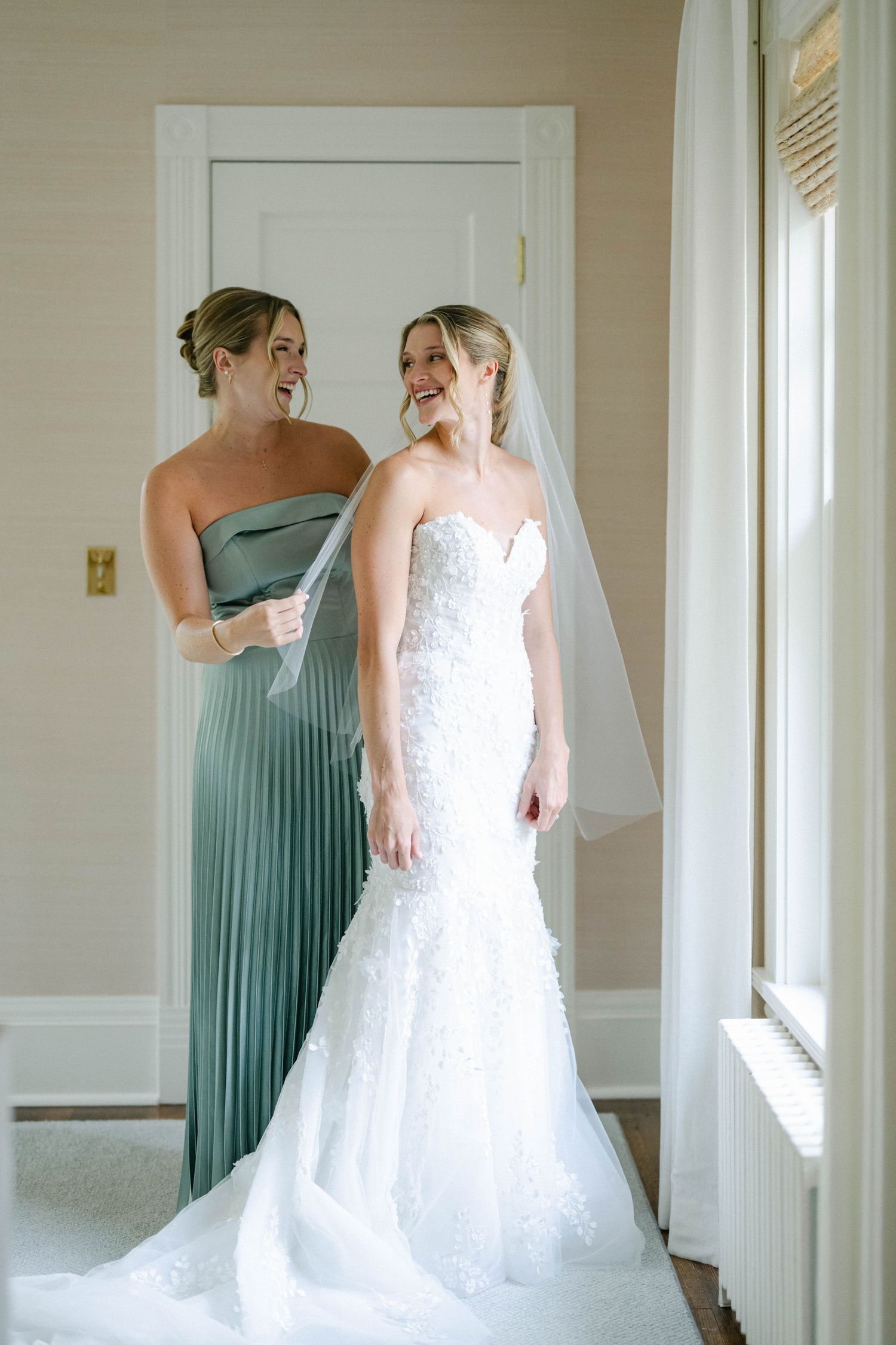 Two women share a joyful moment in a sunlit room, one wearing a sage green pleated gown and one in a white lace gown. - Lake Forest