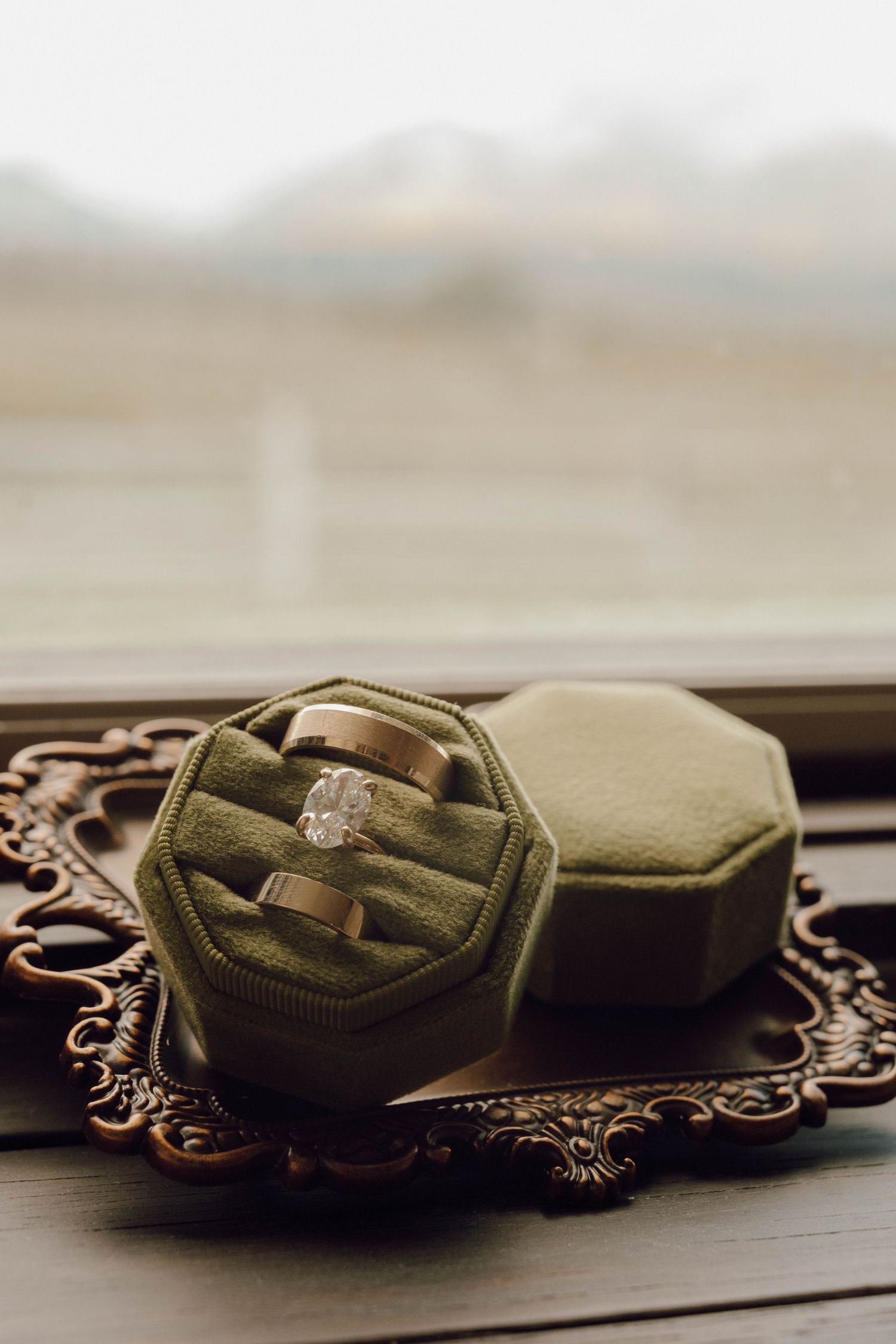 A vintage-style ornate tray displays olive green velvet ring boxes on a wooden surface.