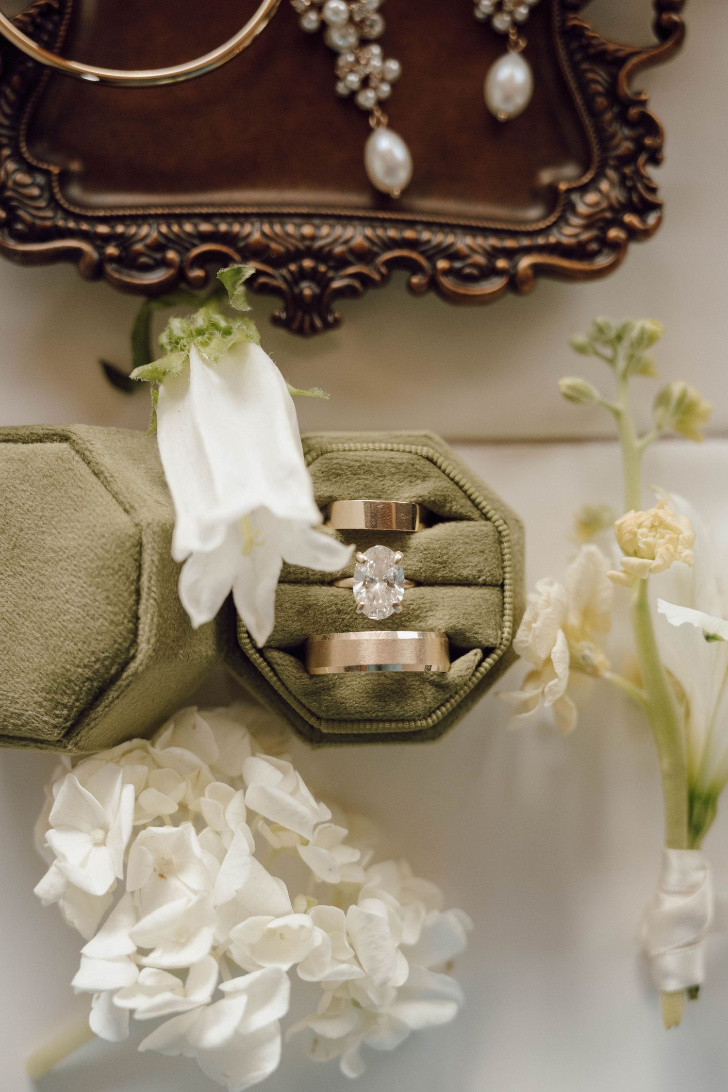 Wedding rings displayed with white flowers and ornate frames on sage green fabric.