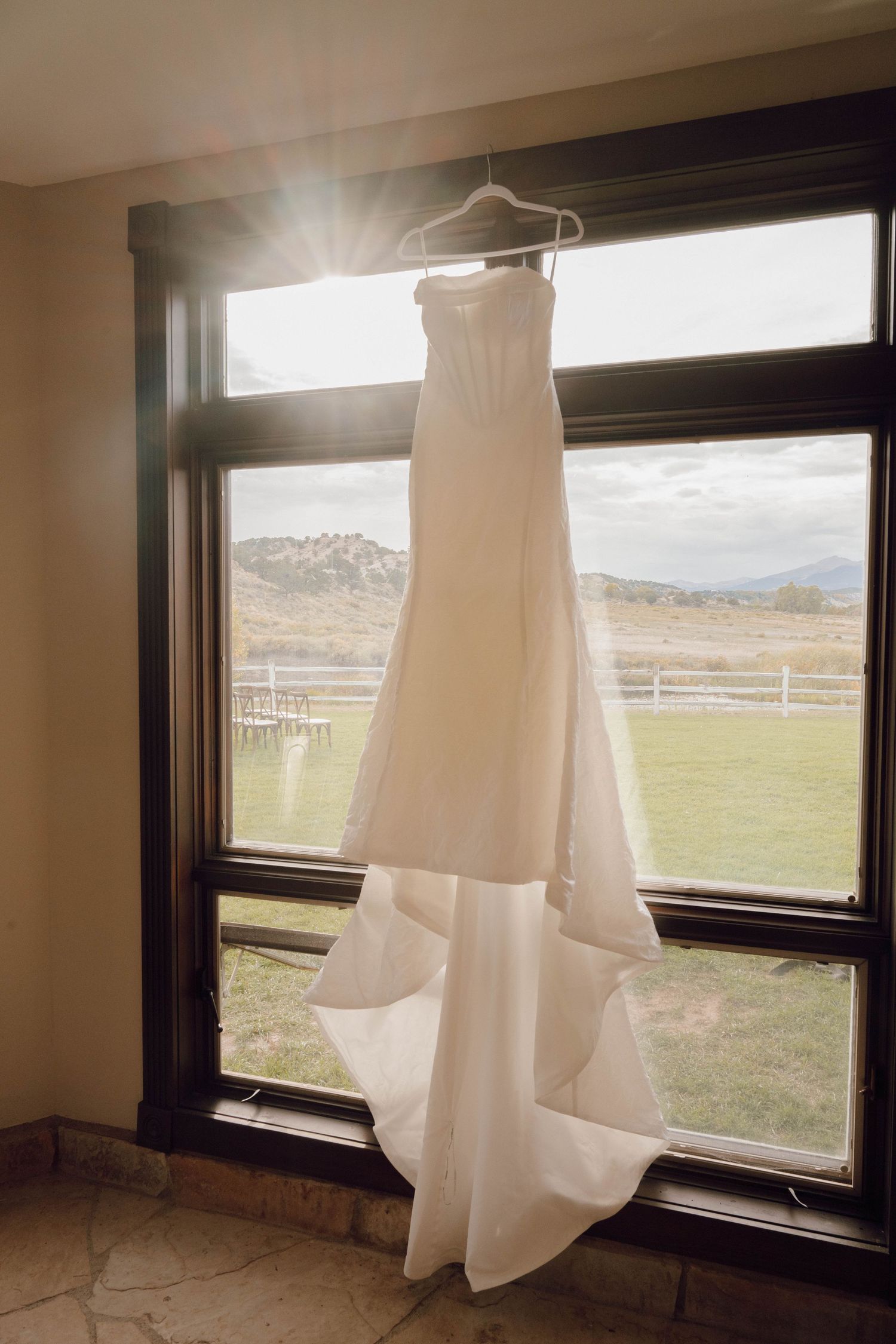 White wedding dress hanging in front of large windows with mountain landscape view.