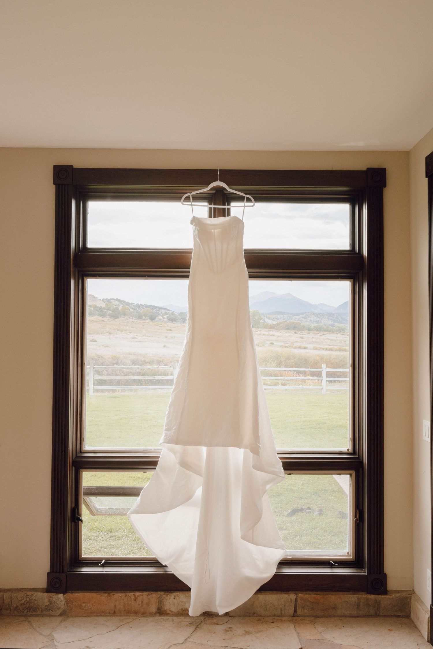 A white wedding dress hangs elegantly in front of a large window with mountain views in the background.