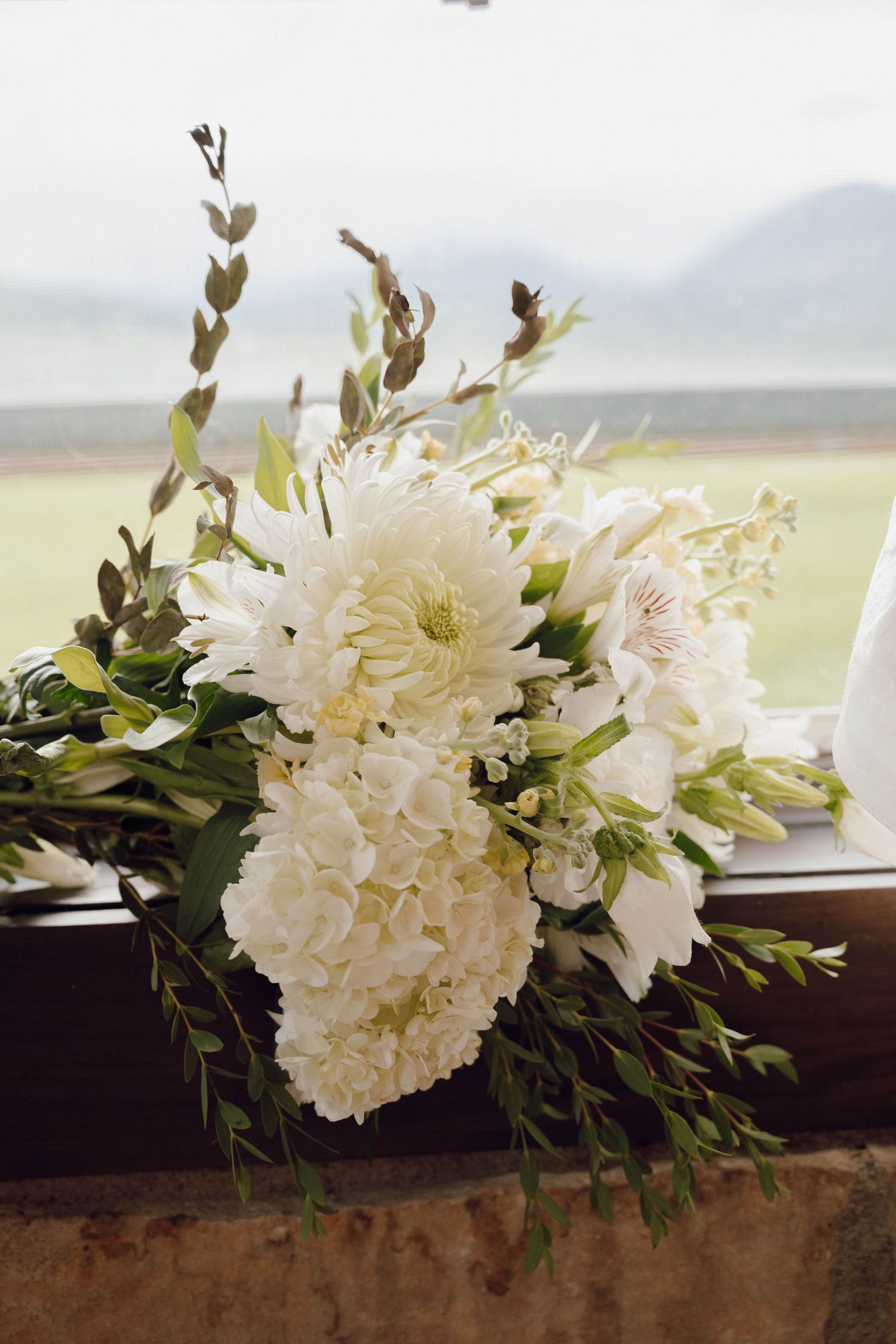 White hydrangeas and floral arrangements line a windowsill with mountain views in the background.